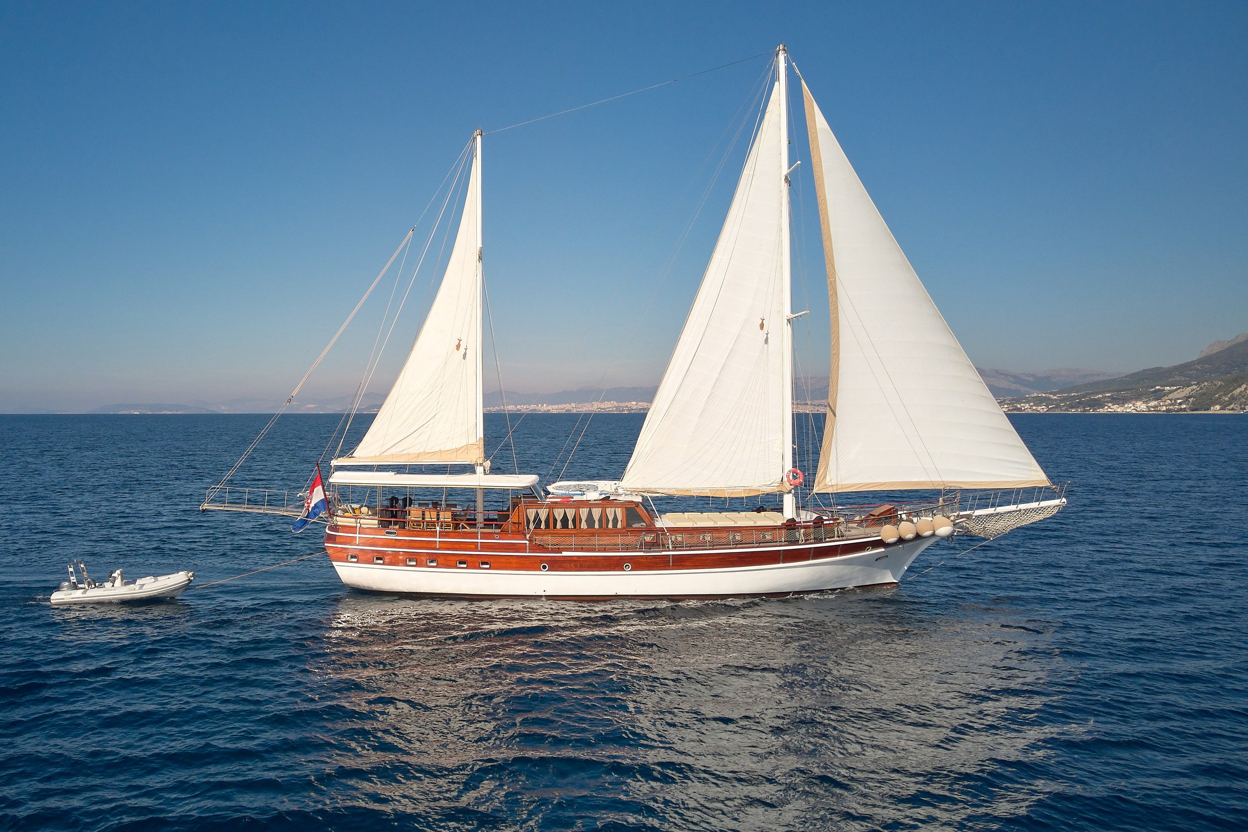 A large sailboat with white sails and a wooden hull sailing on the ocean with a small motorboat nearby, and distant coastline with mountains in the background.