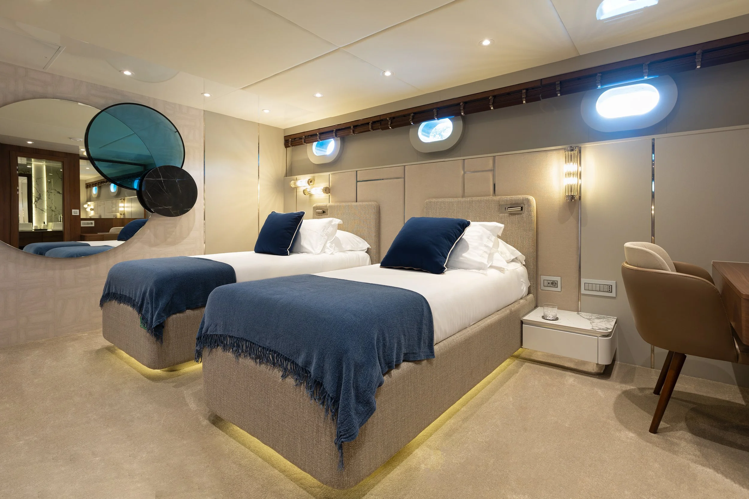 Hotel room with two twin beds featuring beige upholstered headboards, white bedding, navy blue throws, matching navy blue pillows, wall-mounted lights, and a beige armchair at a desk.
