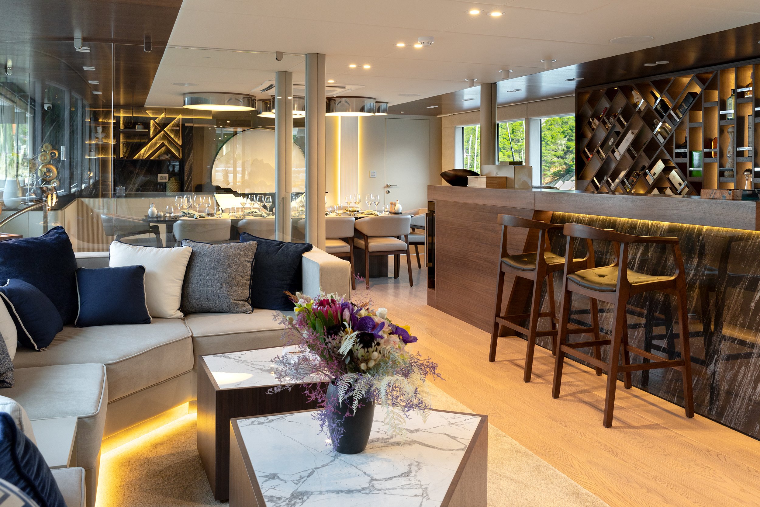 Modern living and dining area with a cream sofa, decorative pillows, a marble-top coffee table with a flower arrangement, a bar counter with barstools, a dining table set with wine glasses, and large windows bringing in natural light.