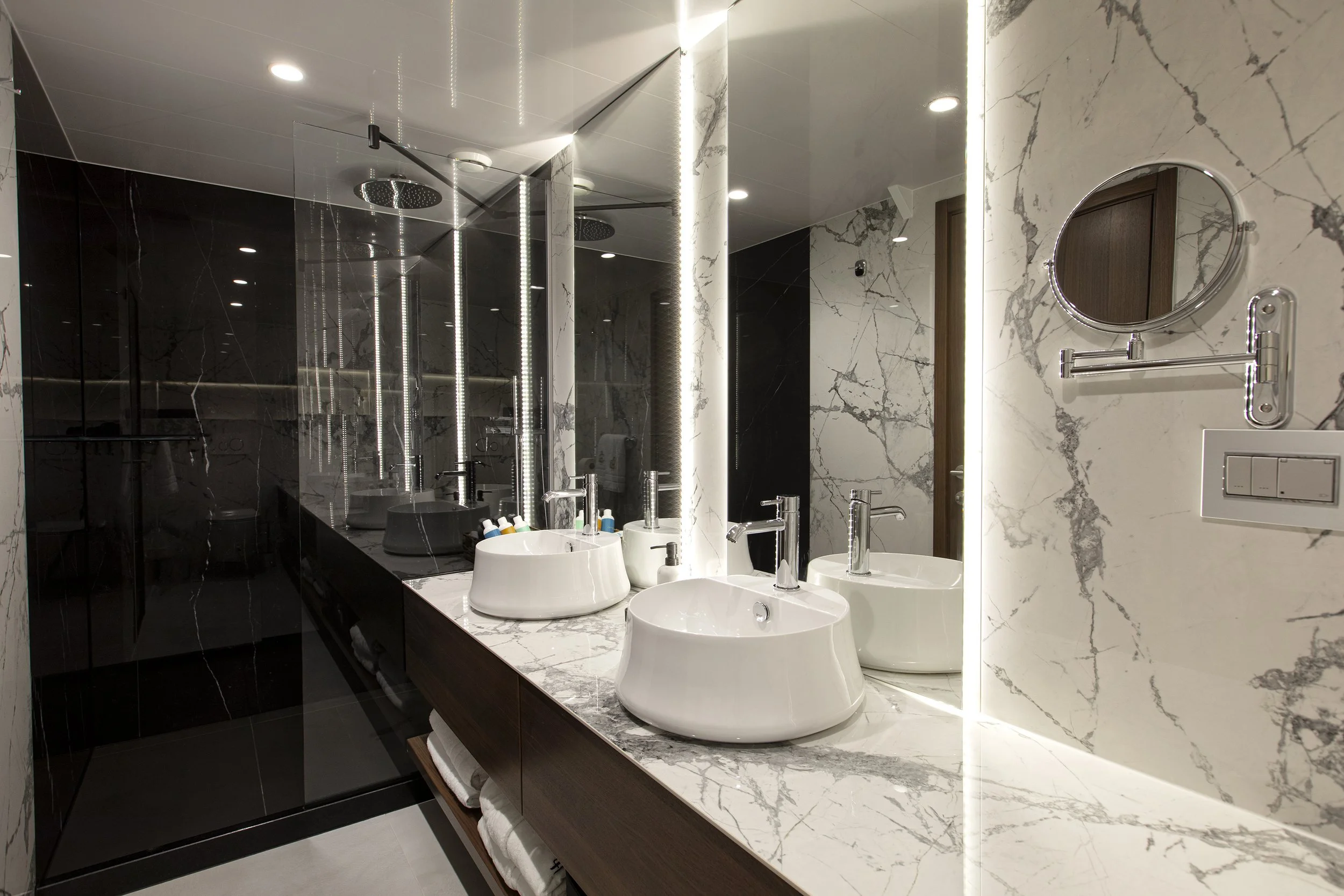 Luxurious bathroom with marble walls and countertop, featuring three black and white vessel sinks with chrome faucets, illuminated mirrors, a black shower area with rainfall showerheads, and a round mirror on the wall.