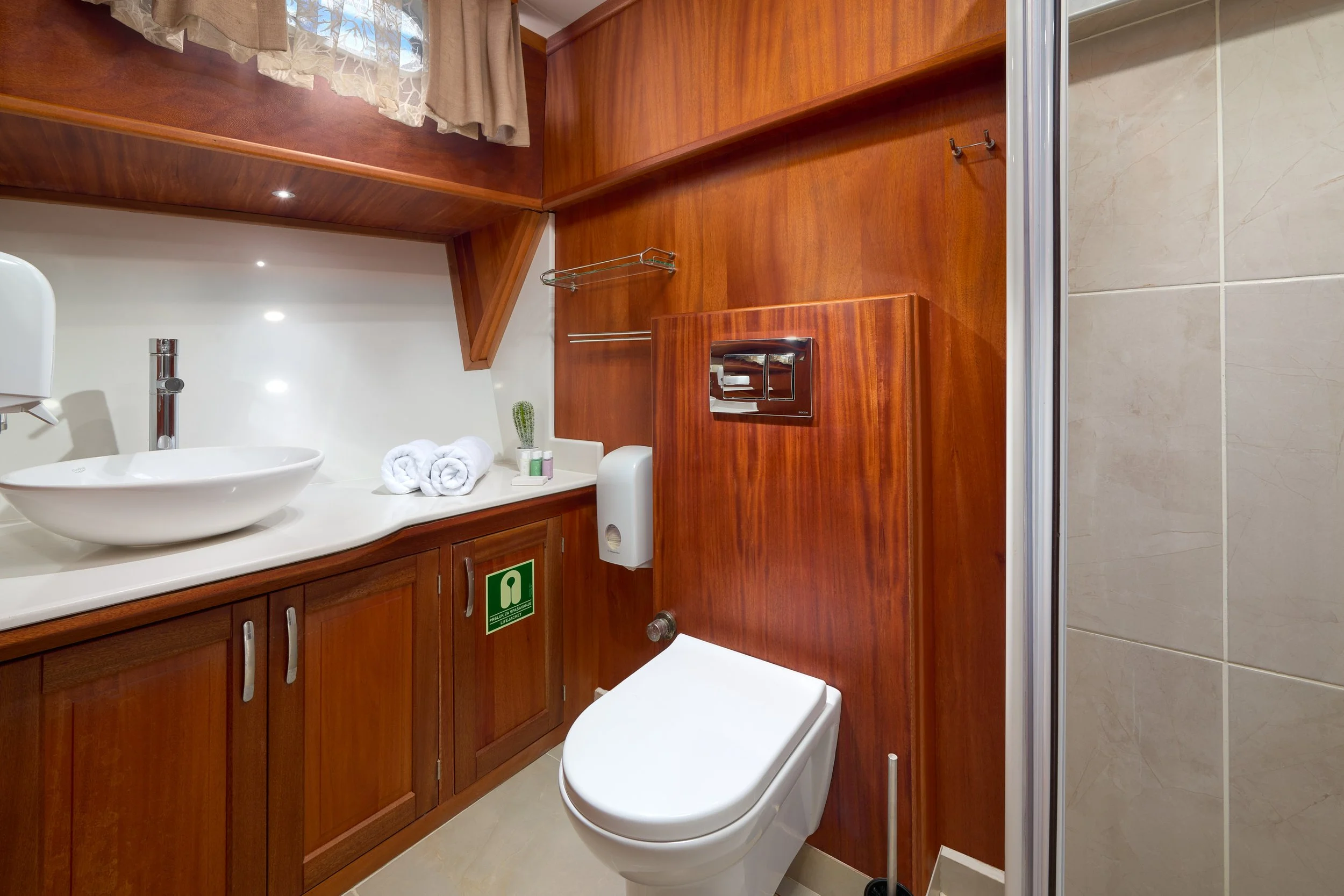 A small bathroom with wood-paneled walls and a white toilet. There is a wooden cabinet with a white countertop, a vessel sink, a small potted cactus, and rolled towels. The bathroom also has a shower enclosure with beige tiles.