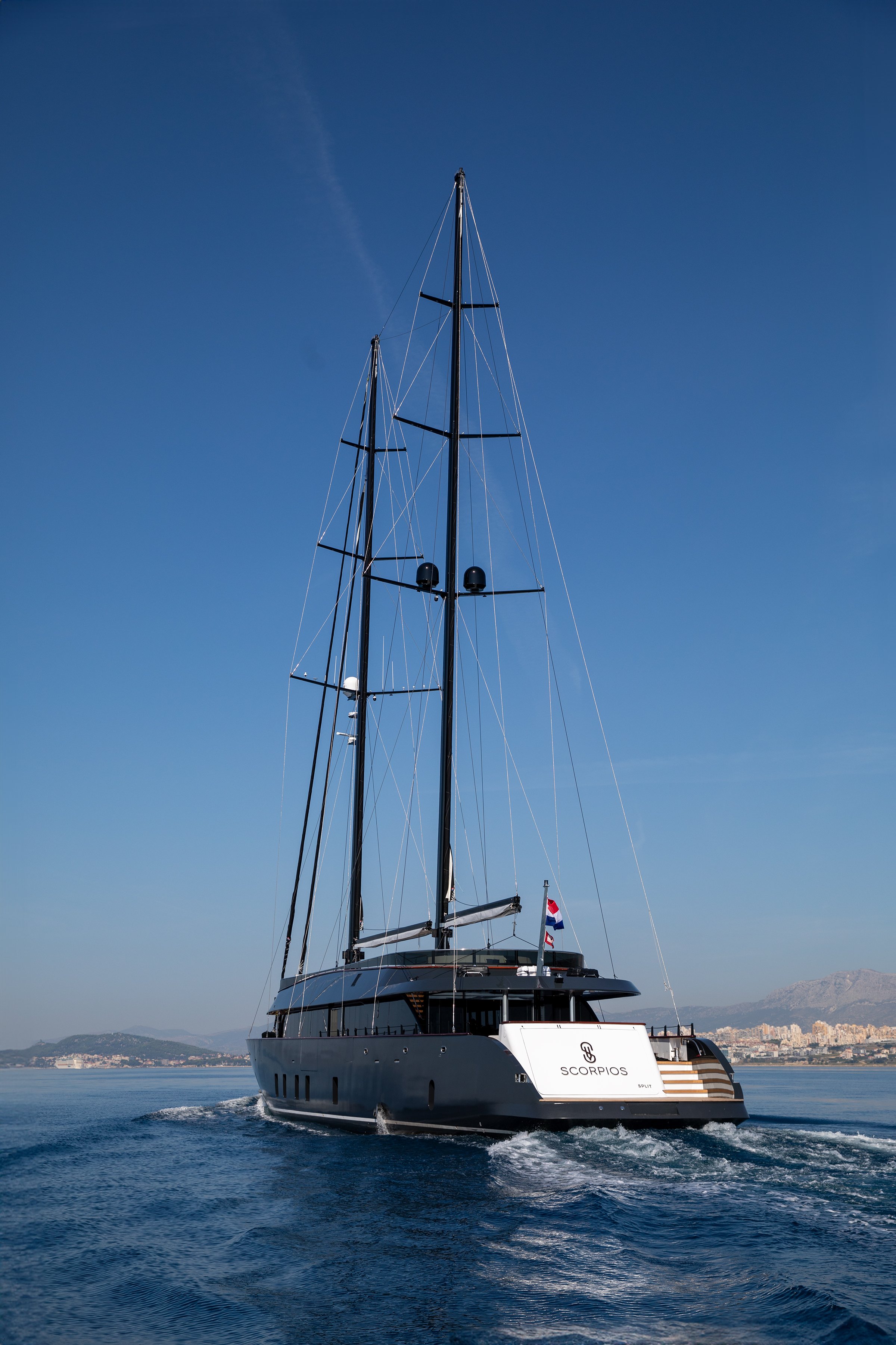 A black luxury sailing yacht named 'Scorpios' cruising in calm blue waters with a city and mountains in the background under a clear sky.