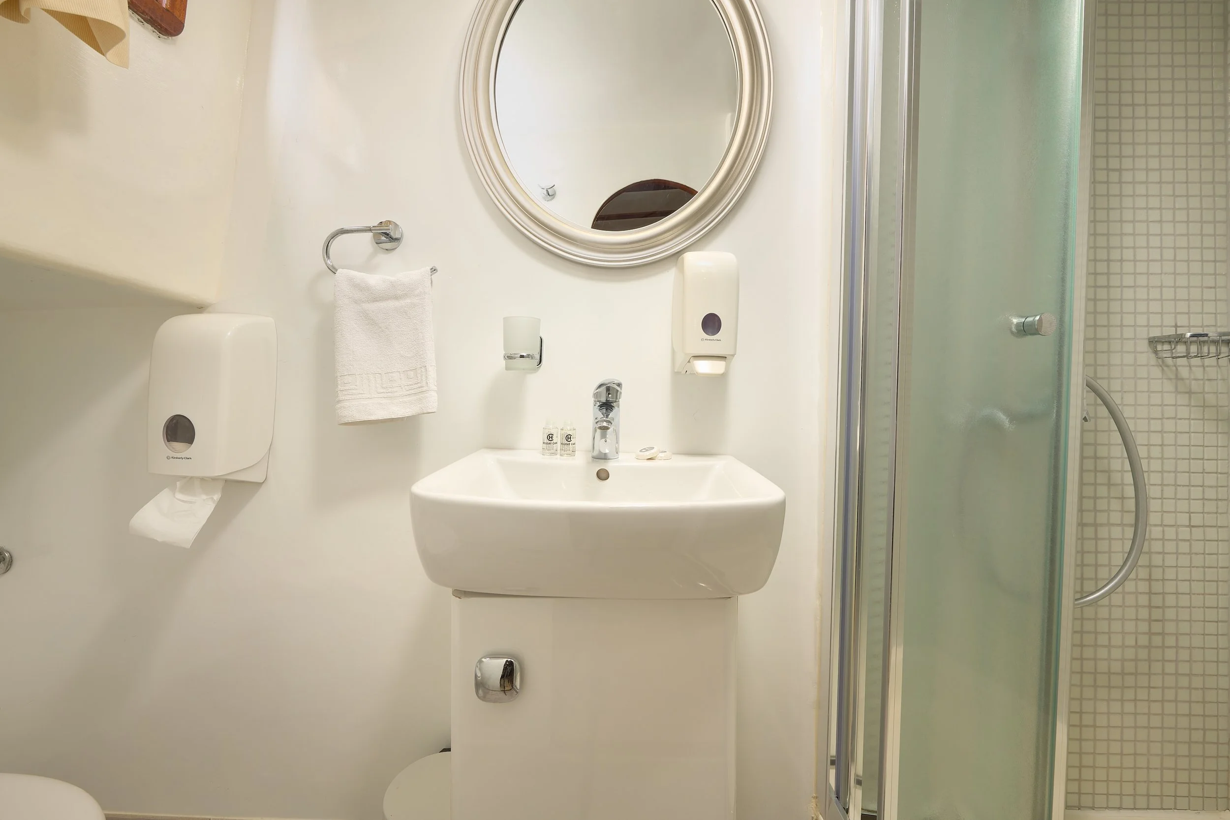 A small hotel bathroom with a white sink, oval mirror, toilet paper dispenser, soap, and a shower with a sliding glass door.