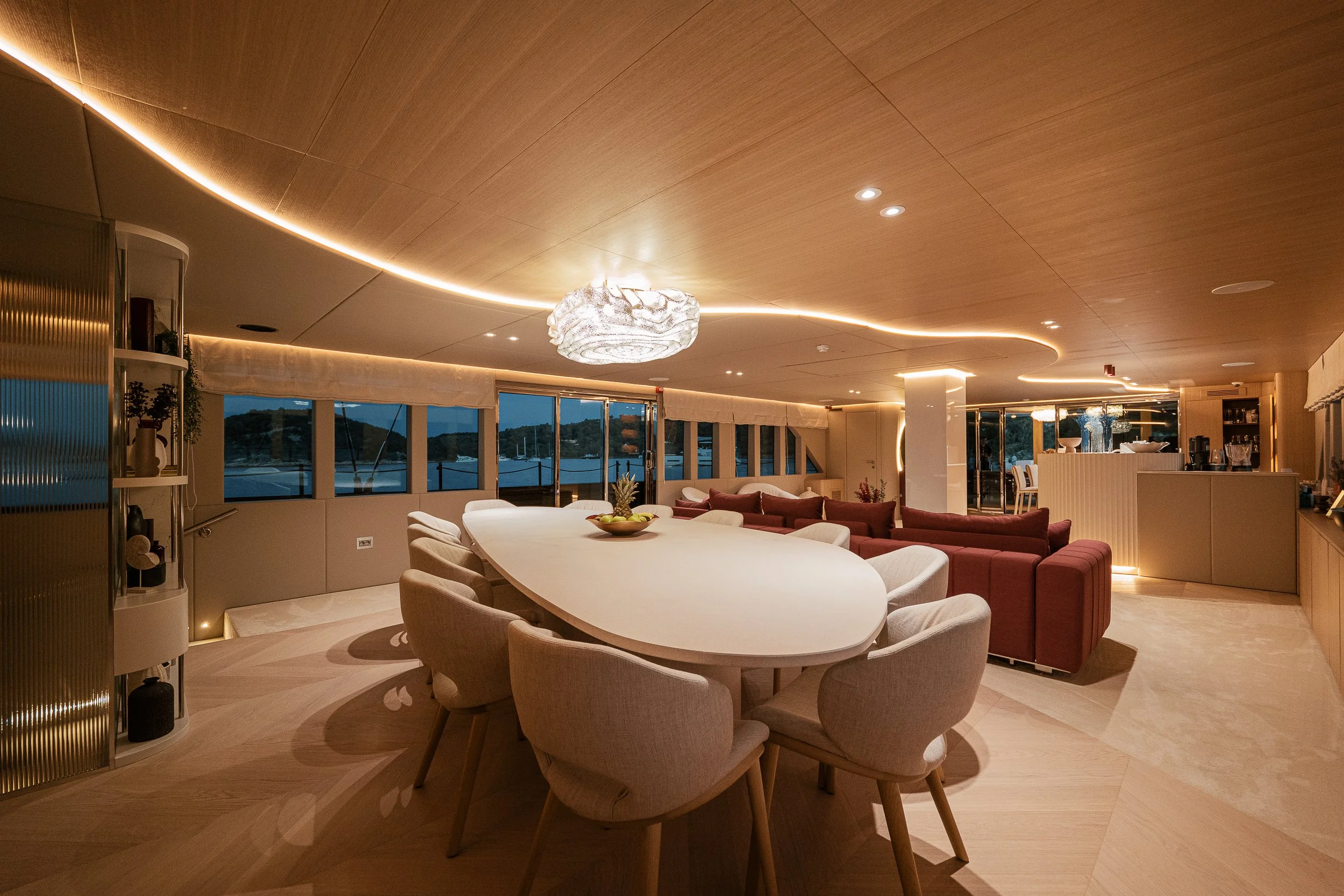 Elegant indoor lounge area with a large oval dining table surrounded by beige chairs, a red sofa, and floor-to-ceiling windows showing an outdoor water view at dusk. Modern lighting fixtures and a bar area are also visible.