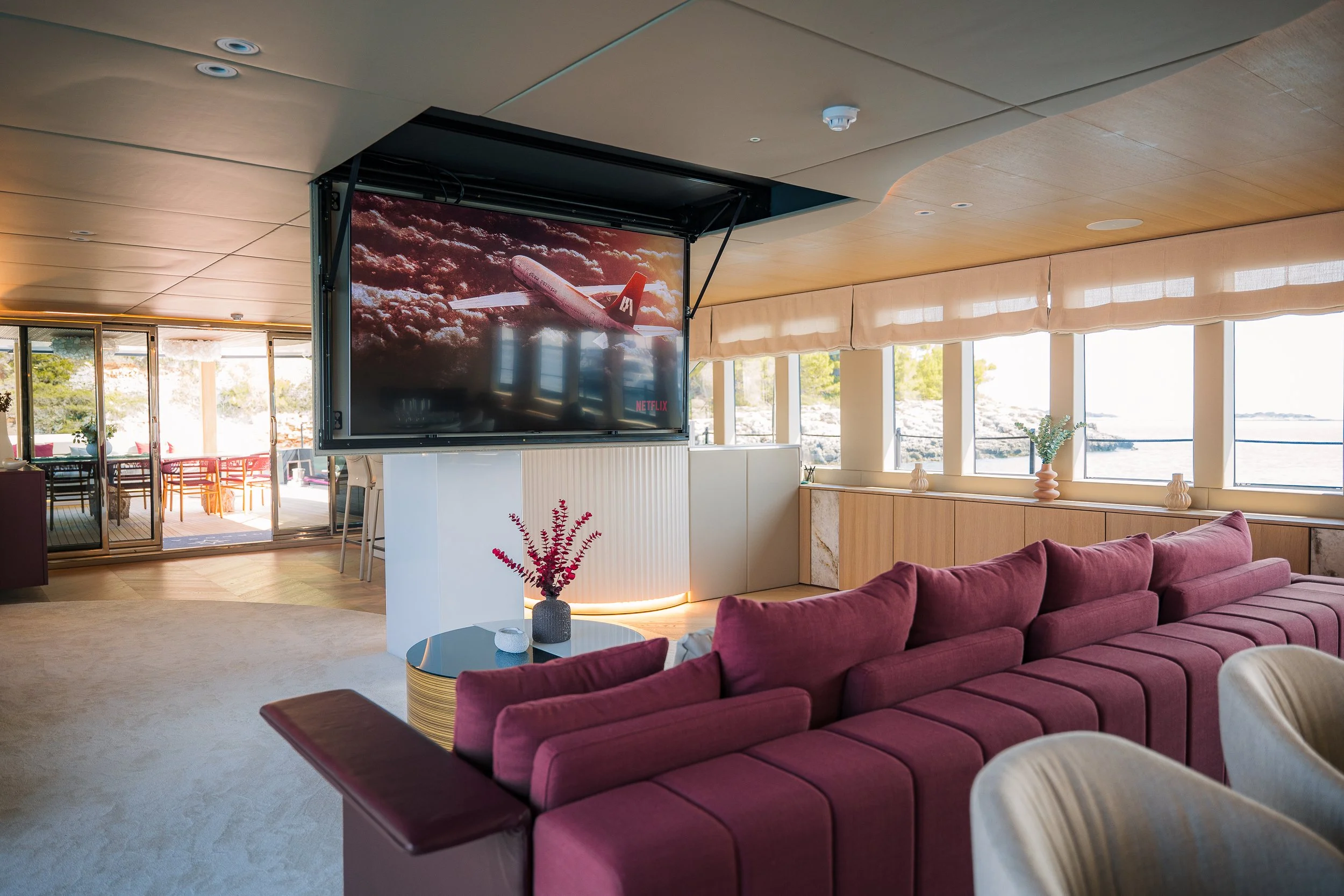Living room with pink sofa, a coffee table with a vase of pink flowers, large windows with a view of water, a television showing an airplane, and sliding glass doors leading to an outdoor patio.