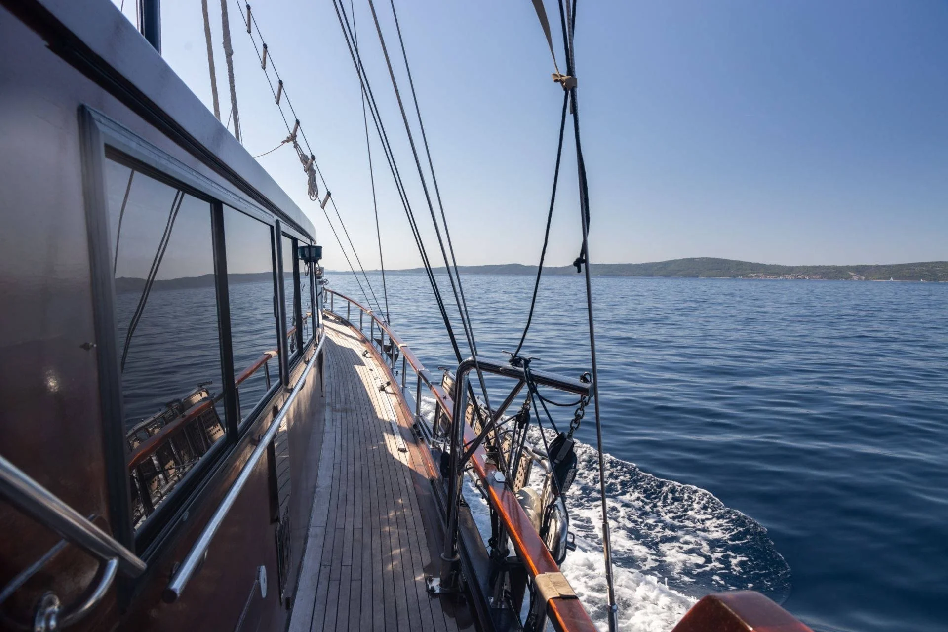 View from the deck of a sailing boat on the open sea with clear blue skies and distant coastline.