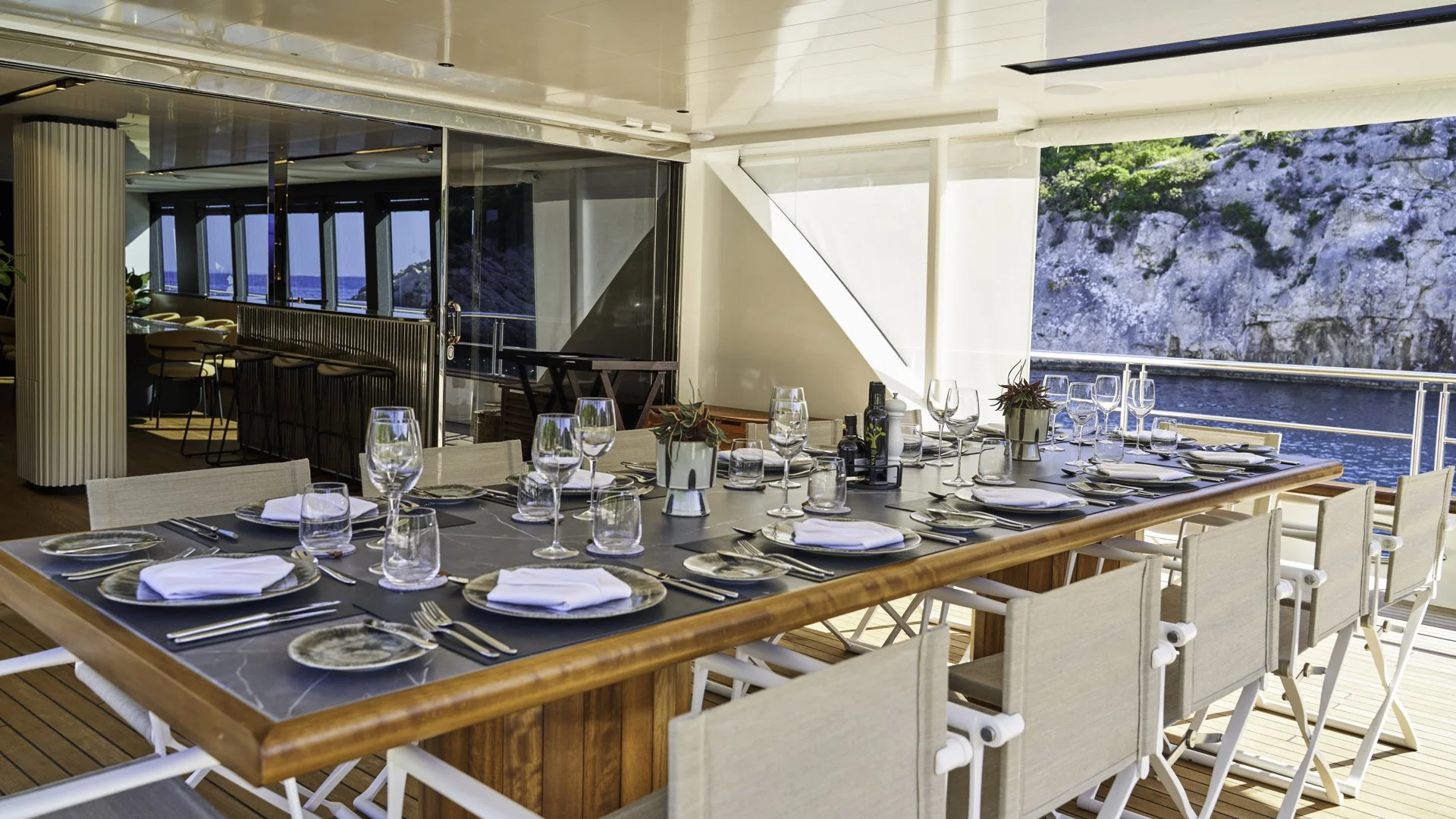 Indoor dining area with a large wooden table set with white napkins, glassware, and plates, overlooking an ocean view with rocky cliffs.