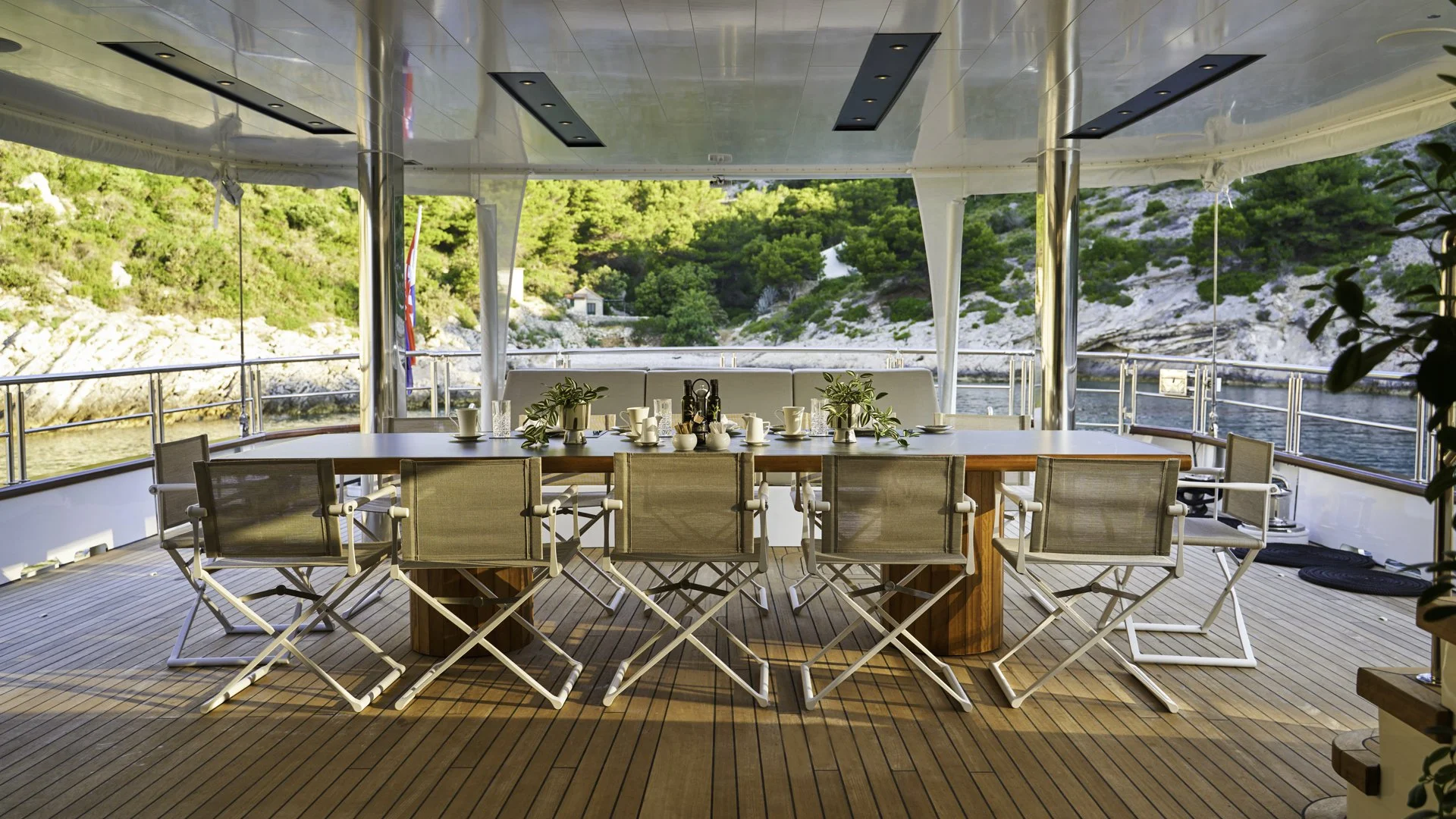A dining table set on the deck of a yacht with seven chairs, potted plants, white cups, and a few bottles, overlooking a waterway with rocky hills and greenery in the background.