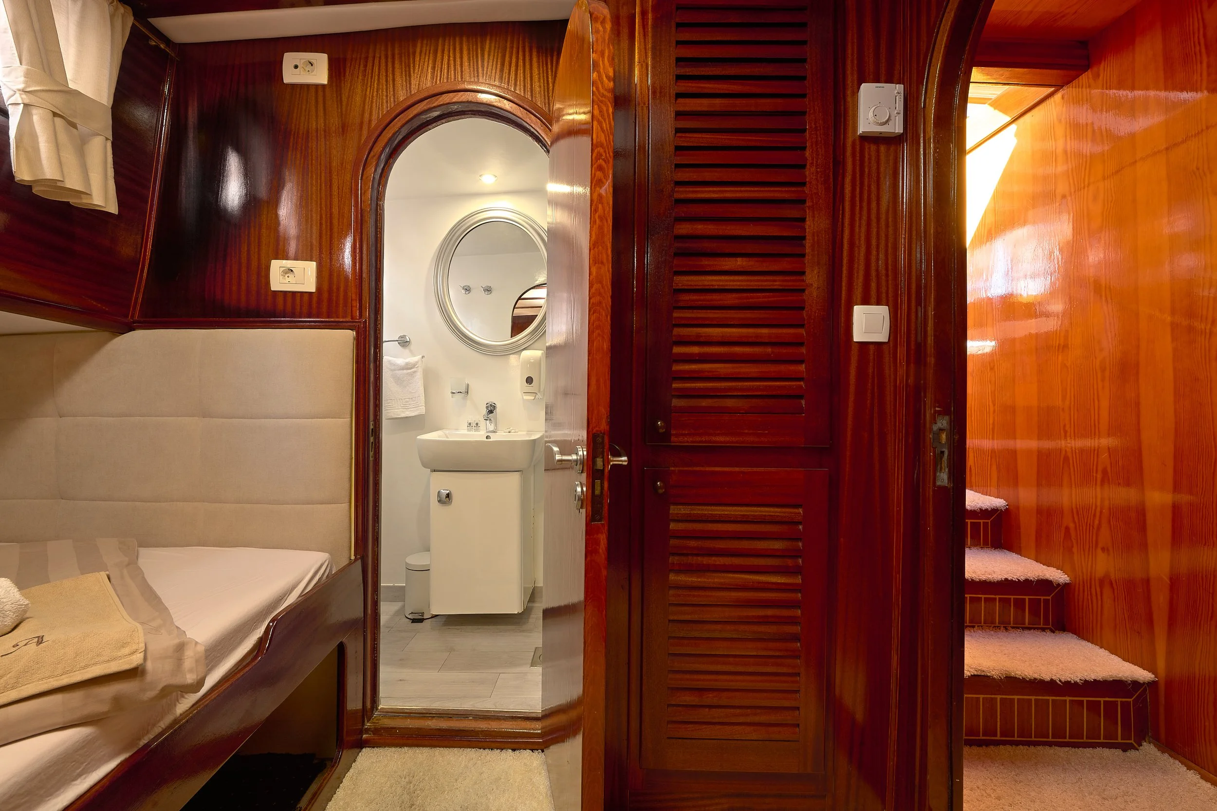 Interior view of a cozy wooden cabin with a small sleeping area on the left, showing a bed with beige linens and a towel, leading to a compact bathroom with a sink, oval mirror, and white walls. To the right, wooden louvered doors cover a closet, and