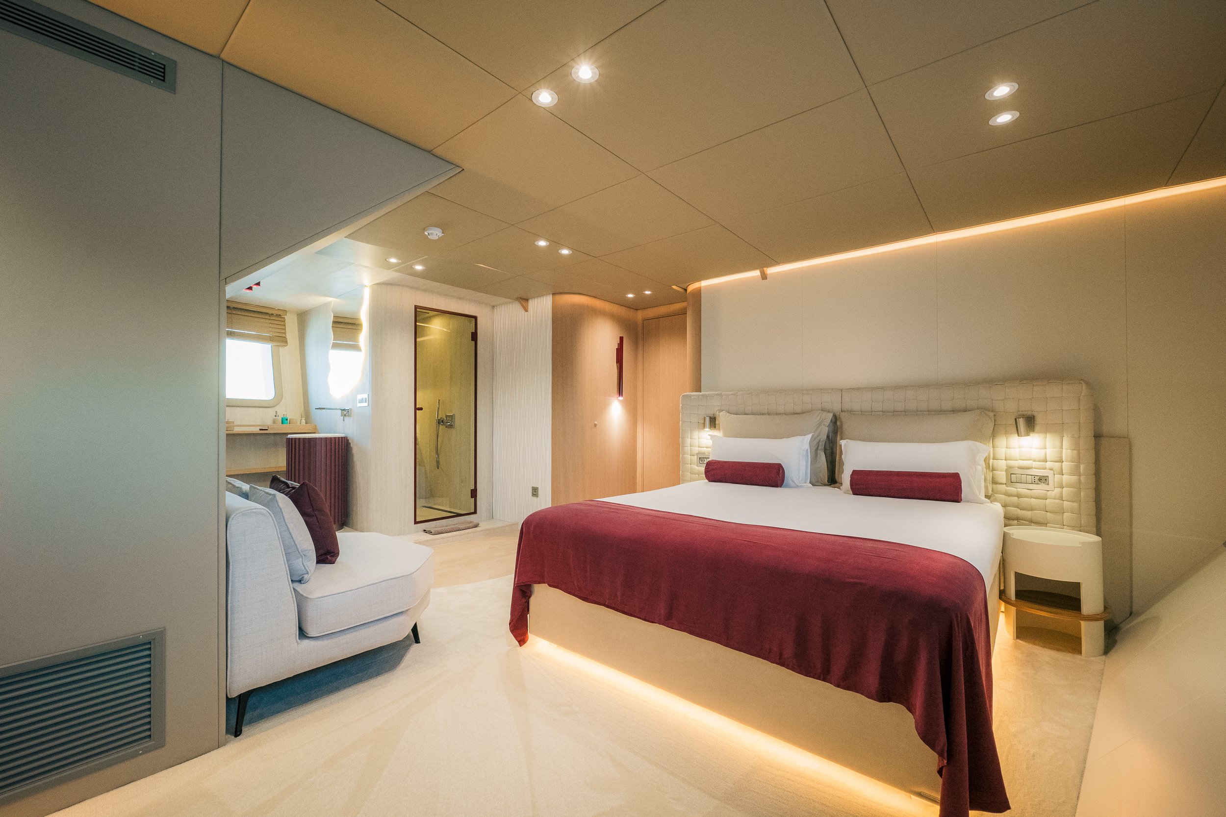 Modern hotel room with a large bed, white headboard, maroon blanket, pillows, a small sofa with pillows, and a shower area in the background.