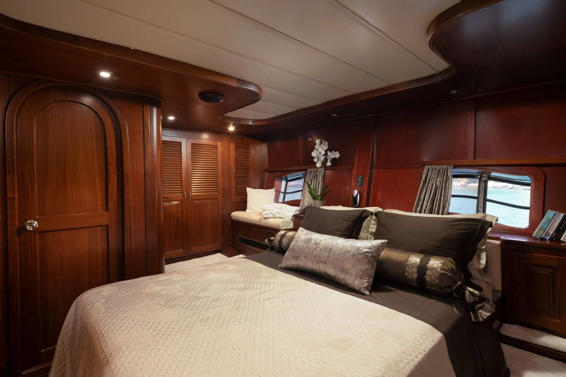 Luxurious yacht bedroom interior with wood paneling, a neatly made bed with decorative pillows, and a window overlooking water.