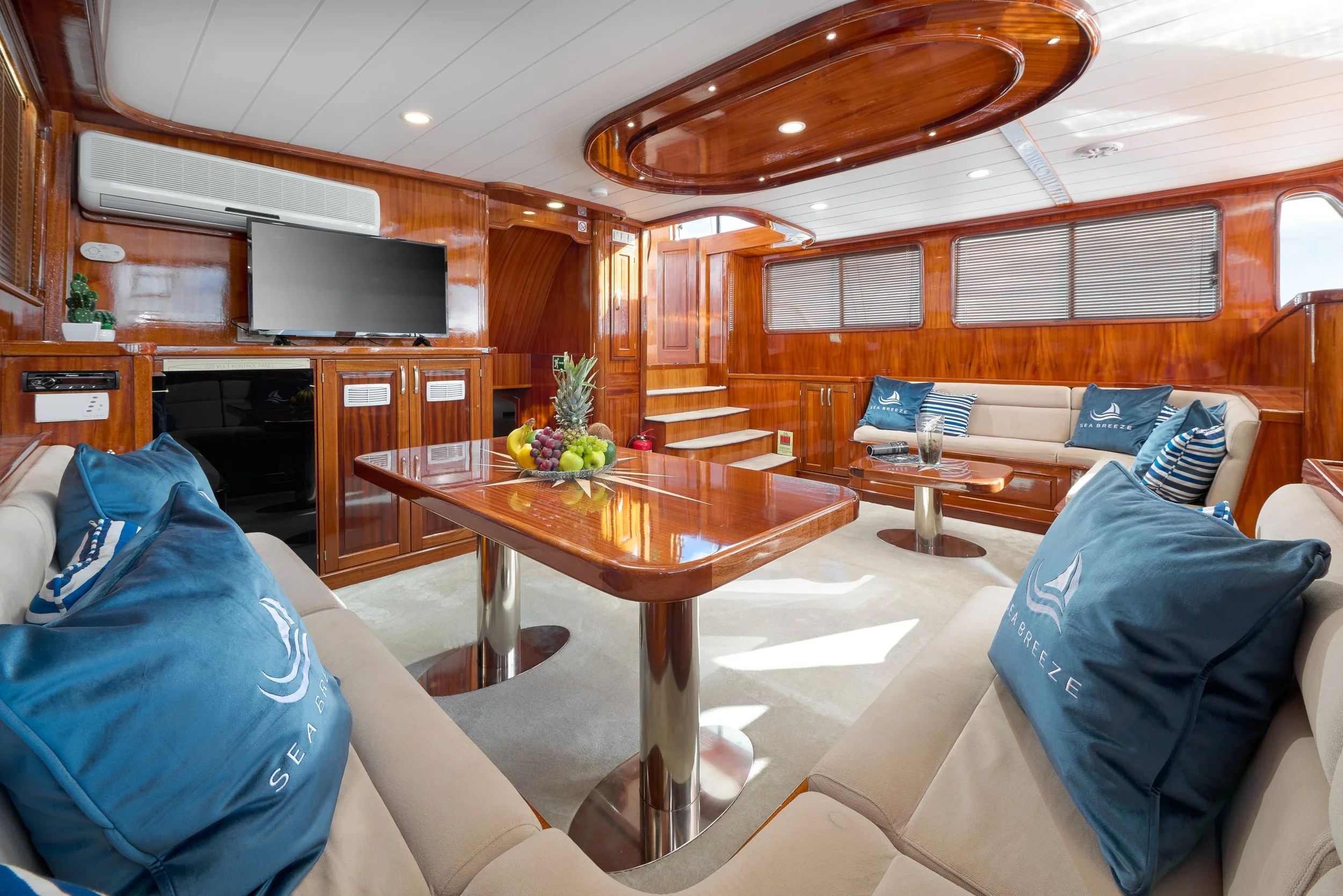 Luxurious wooden-paneled yacht interior with beige sofas adorned with blue pillows, a wooden dining table with a fruit centerpiece, a wall-mounted TV, and window blinds.