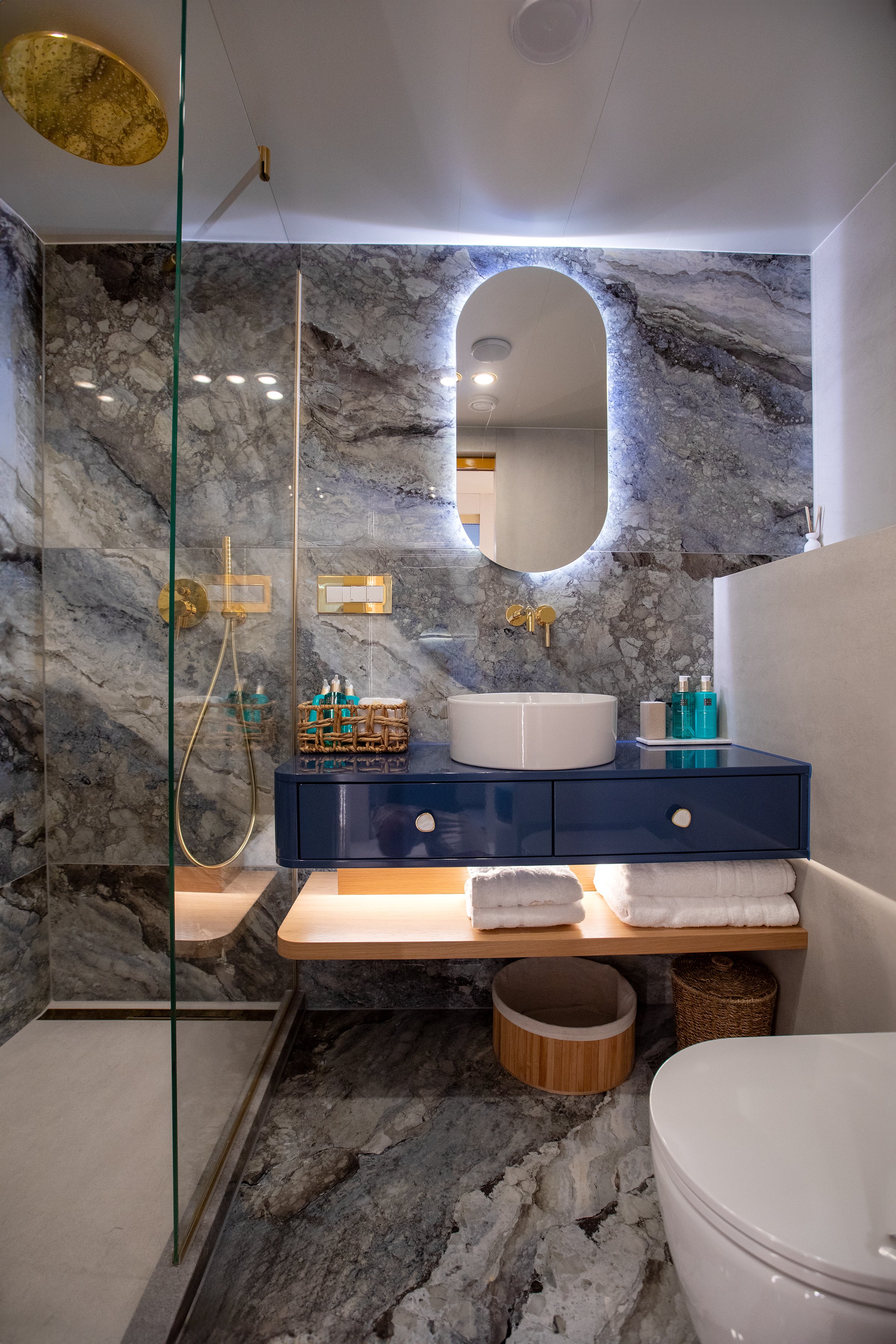 Modern bathroom with marble walls and floor, a blue vanity with a round white sink, a mirror with backlighting, a glass shower with gold fixtures, and neatly folded towels.