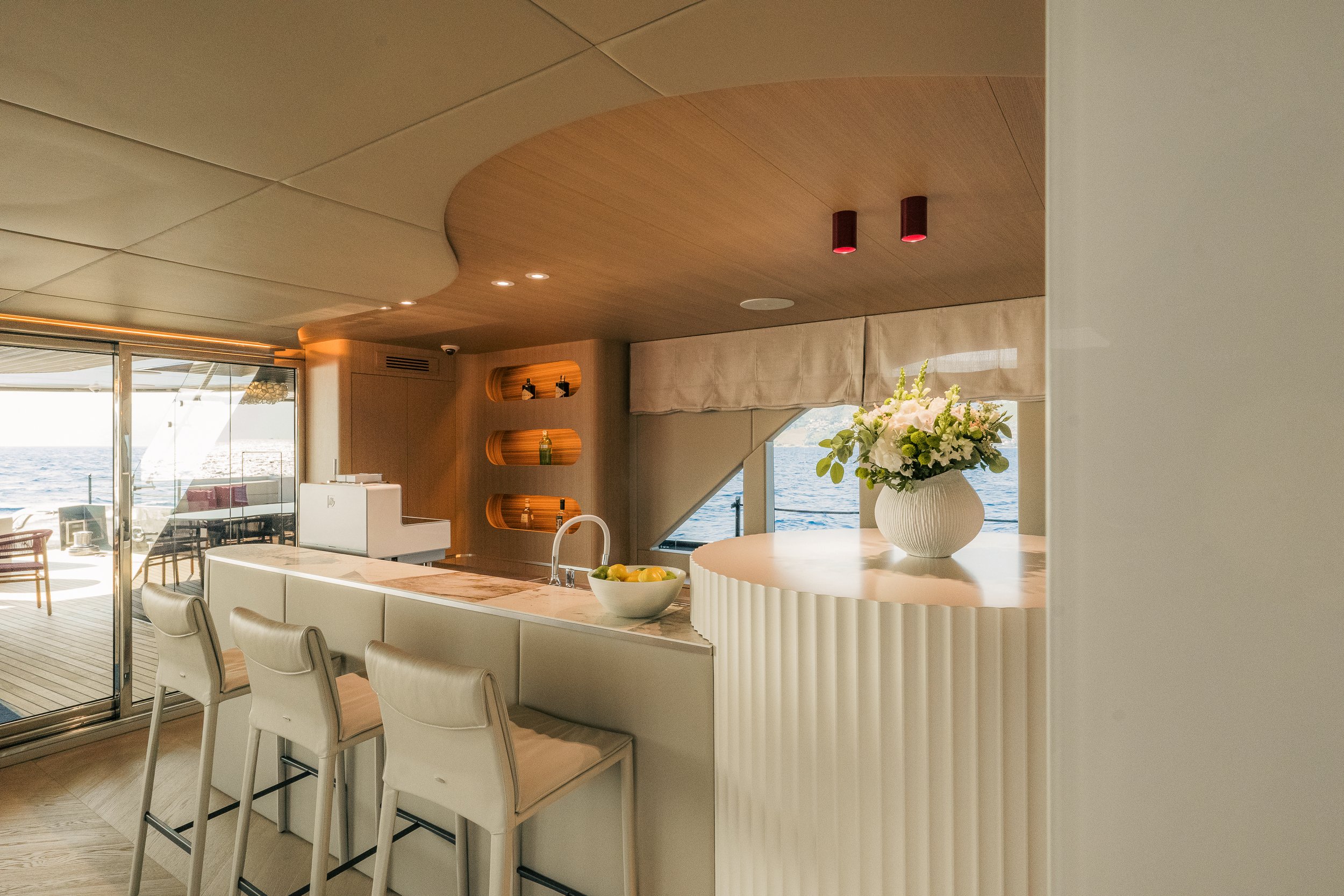 Interior of a modern yacht kitchen with a curved white island, a floral vase, and a bowl of fruit, with ocean views outside the windows.