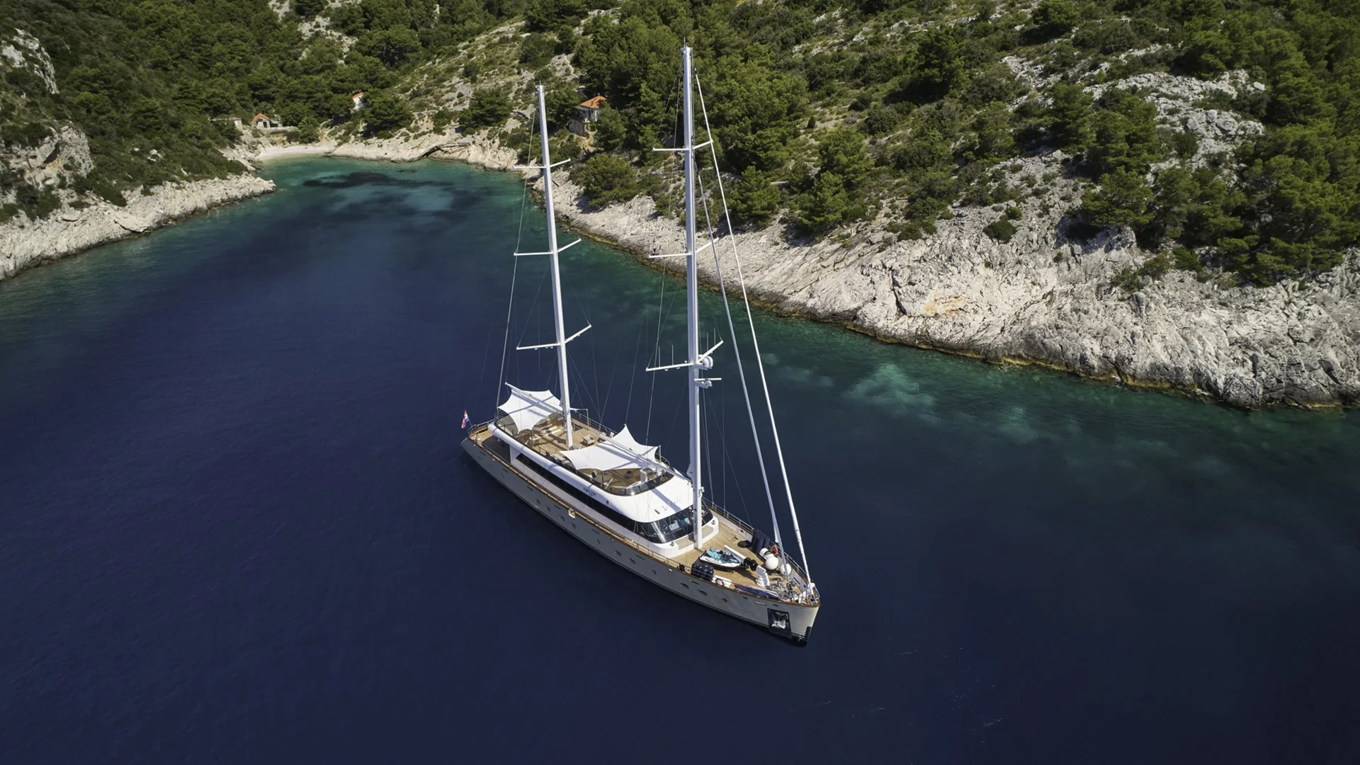 An aerial view of a white sailing yacht anchored in a calm, blue sea near a rocky green coastline with trees and small structures.