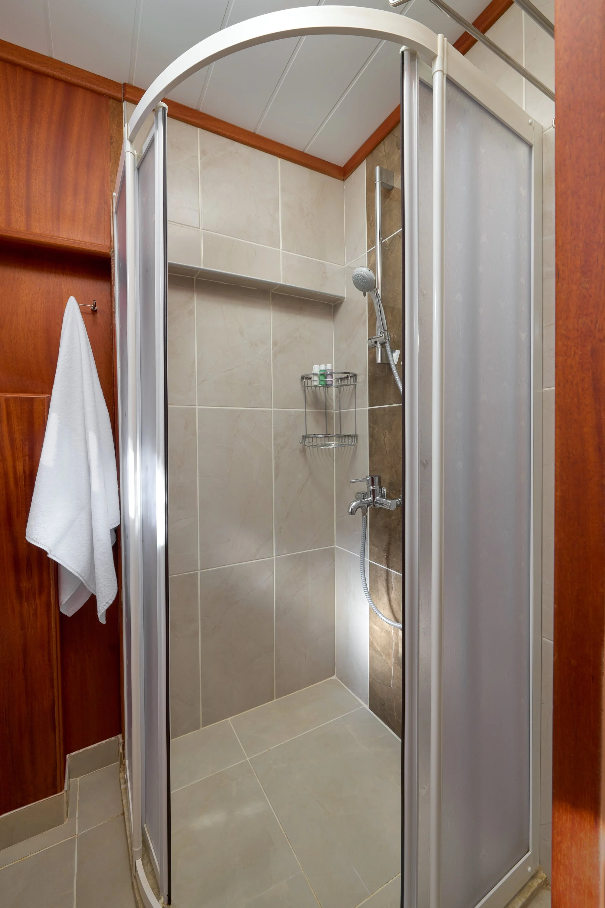 Small shower stall with beige and brown tiled walls, a sliding frosted glass door, a handheld showerhead, and a metal corner caddy with toiletries. There is a white towel hanging on a wooden wall hook outside the shower.