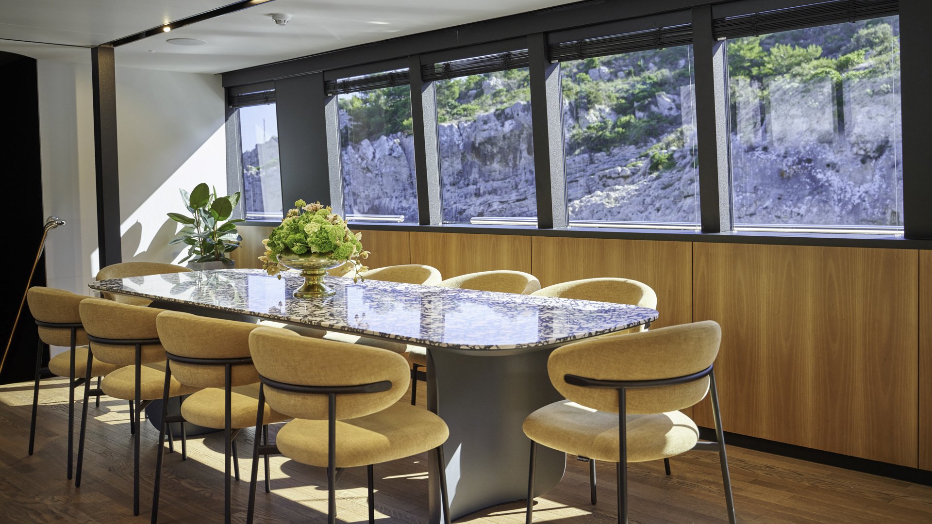 Modern dining room with a marble-topped table, surrounded by tan upholstered chairs. Large windows reveal a rocky hillside and greenery outside, with a potted plant and floral arrangement on the table.