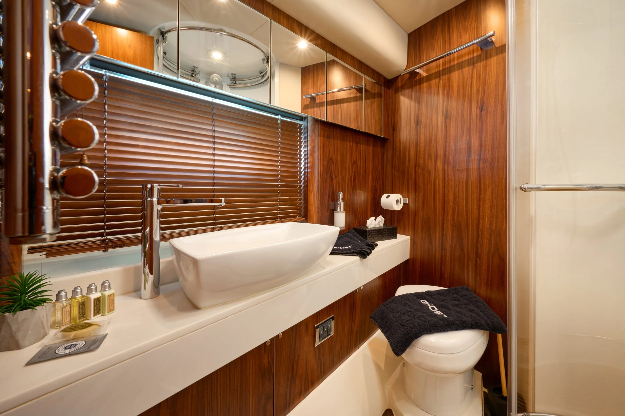 Modern bathroom with wood-paneled wall, white countertop with vessel sink, mirror, blinds on window, and toilet with black towel draped over it.