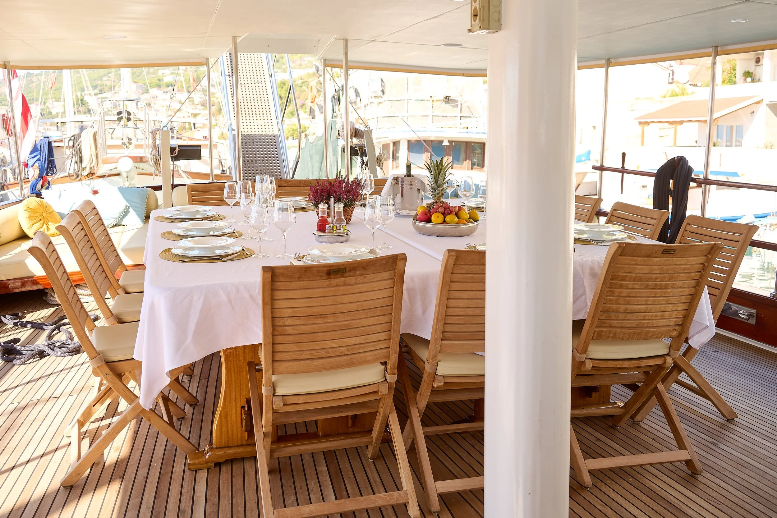 A boat deck set for a meal with a large round table covered in a white tablecloth, surrounded by wooden chairs with cushions, with boats and water visible in the background.