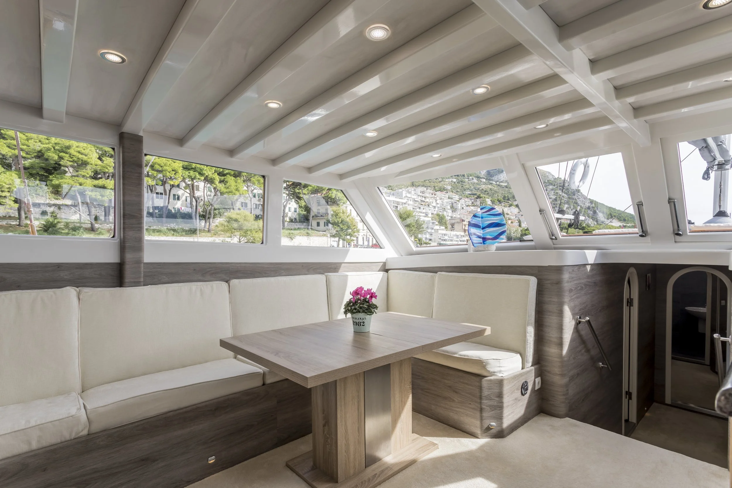 Interior of a boat with a cream-colored curved bench seating arrangement, a wooden dining table with a pink flowering plant, large windows showing a hillside neighborhood, and a ceiling with recessed lighting.