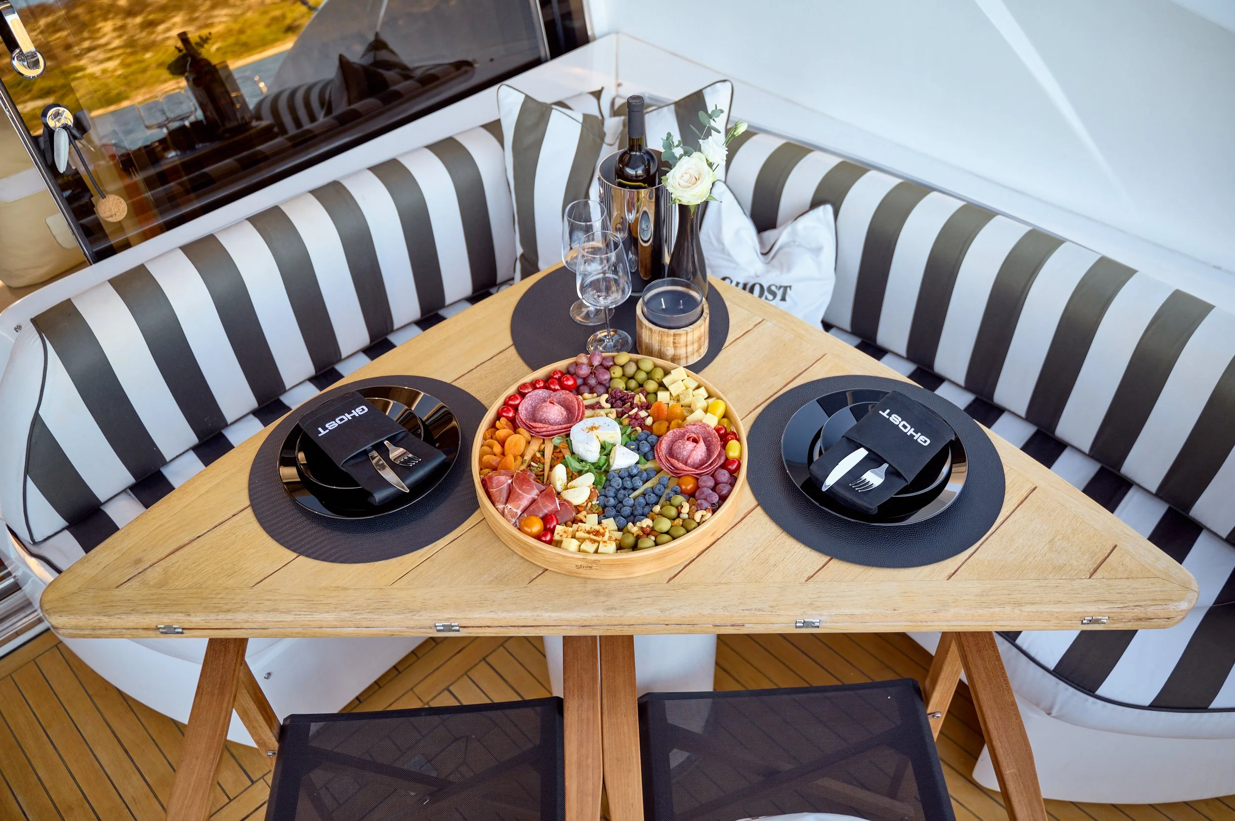 A triangular wooden outdoor dining table set for two, with black round placemats, black plates with cutlery wrapped in black napkins labeled 'GHOST', a large charcuterie and fruit platter, a wine bottle with glasses, a candle, and a white flower in a