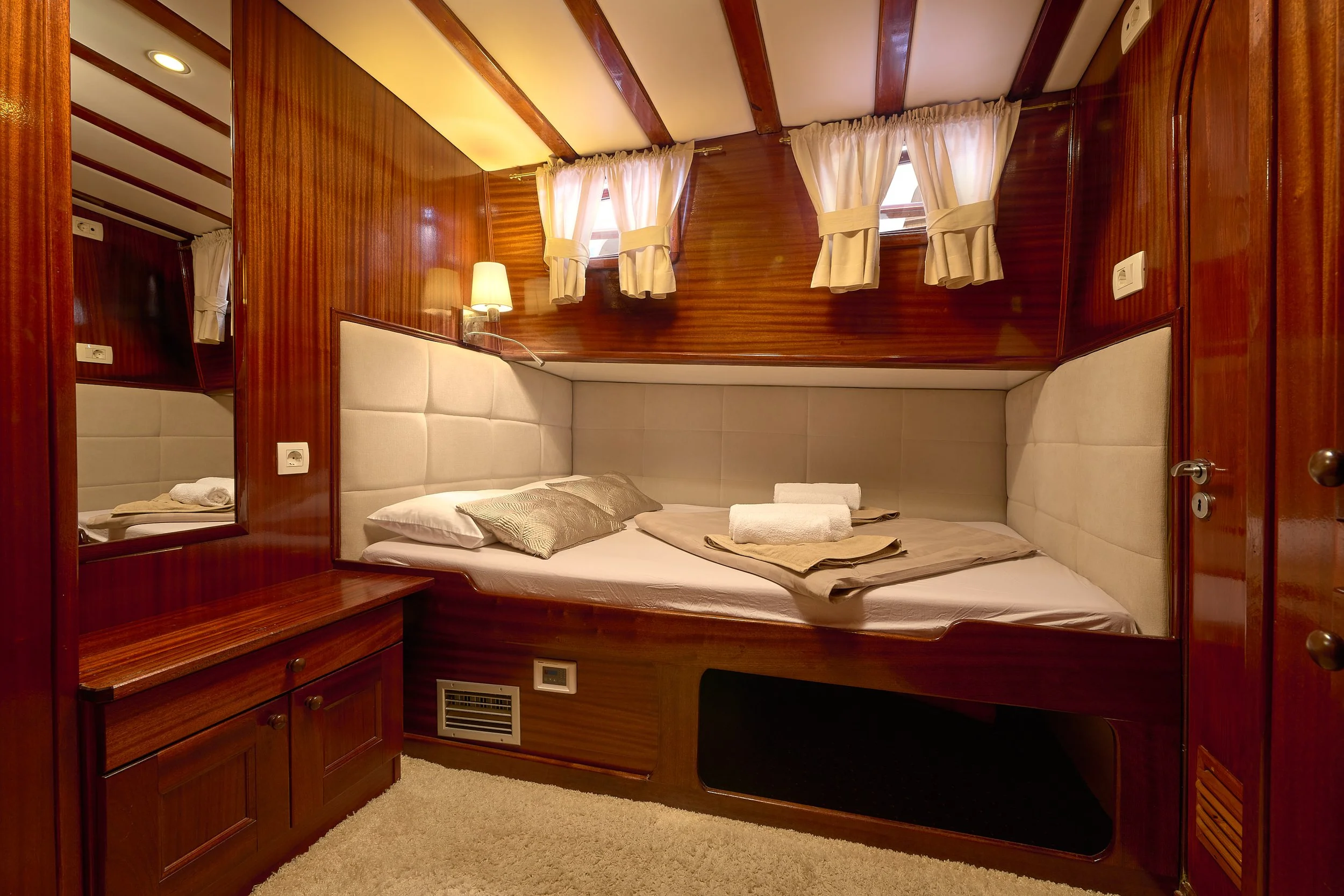 Small bedroom with wooden panel walls, a bed with beige cushions, two rolled towels, and windows with white curtains.