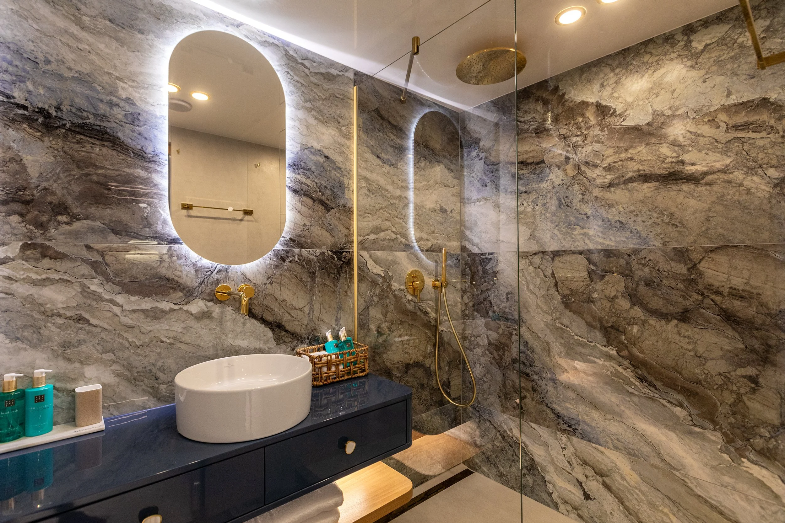 Modern bathroom with marble walls, a large backlit mirror, a vessel sink, gold fixtures, and a glass-enclosed shower area.