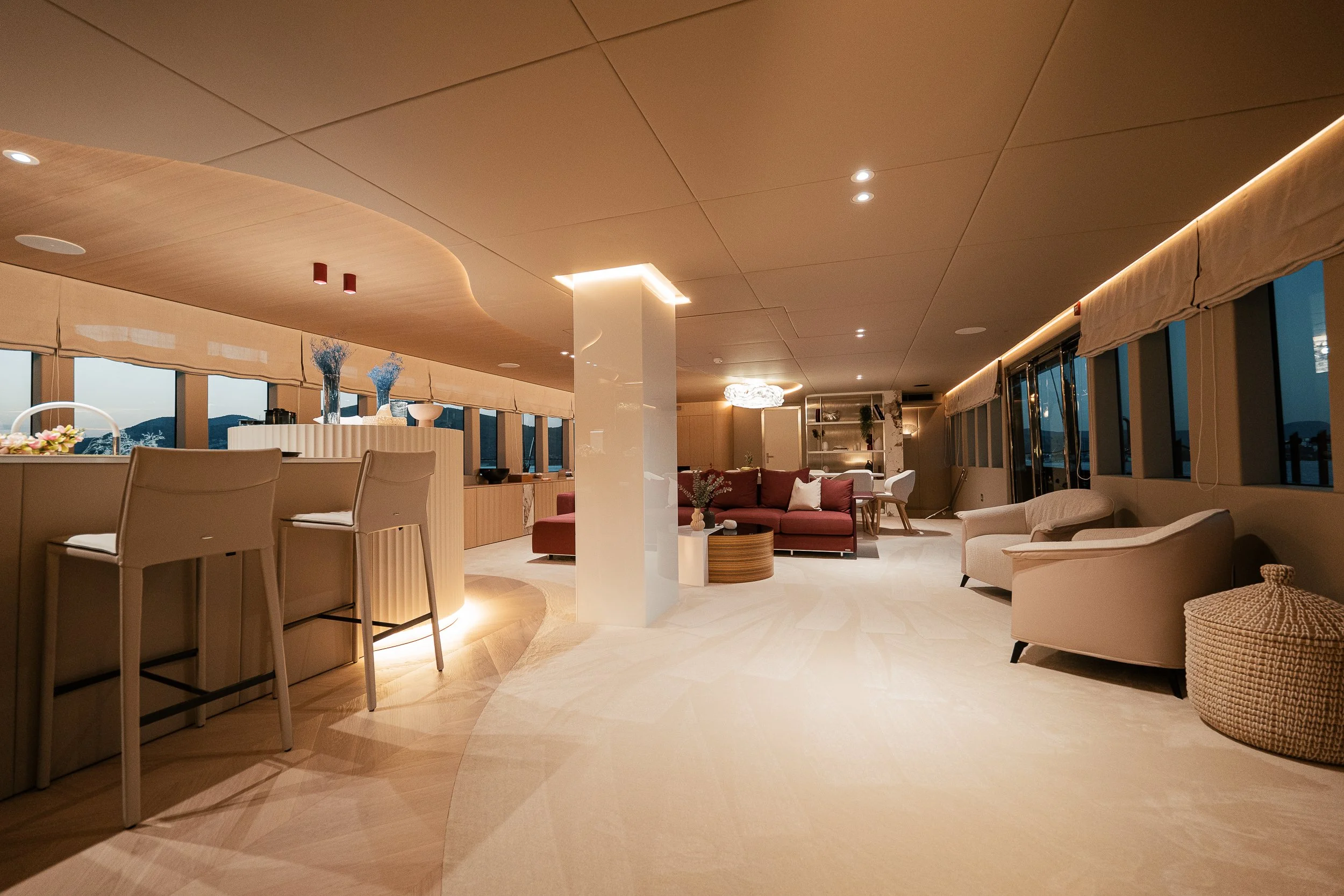 Spacious modern living room with a bar area, high chairs, a maroon sofa, beige armchairs, and large windows with a sunset view.