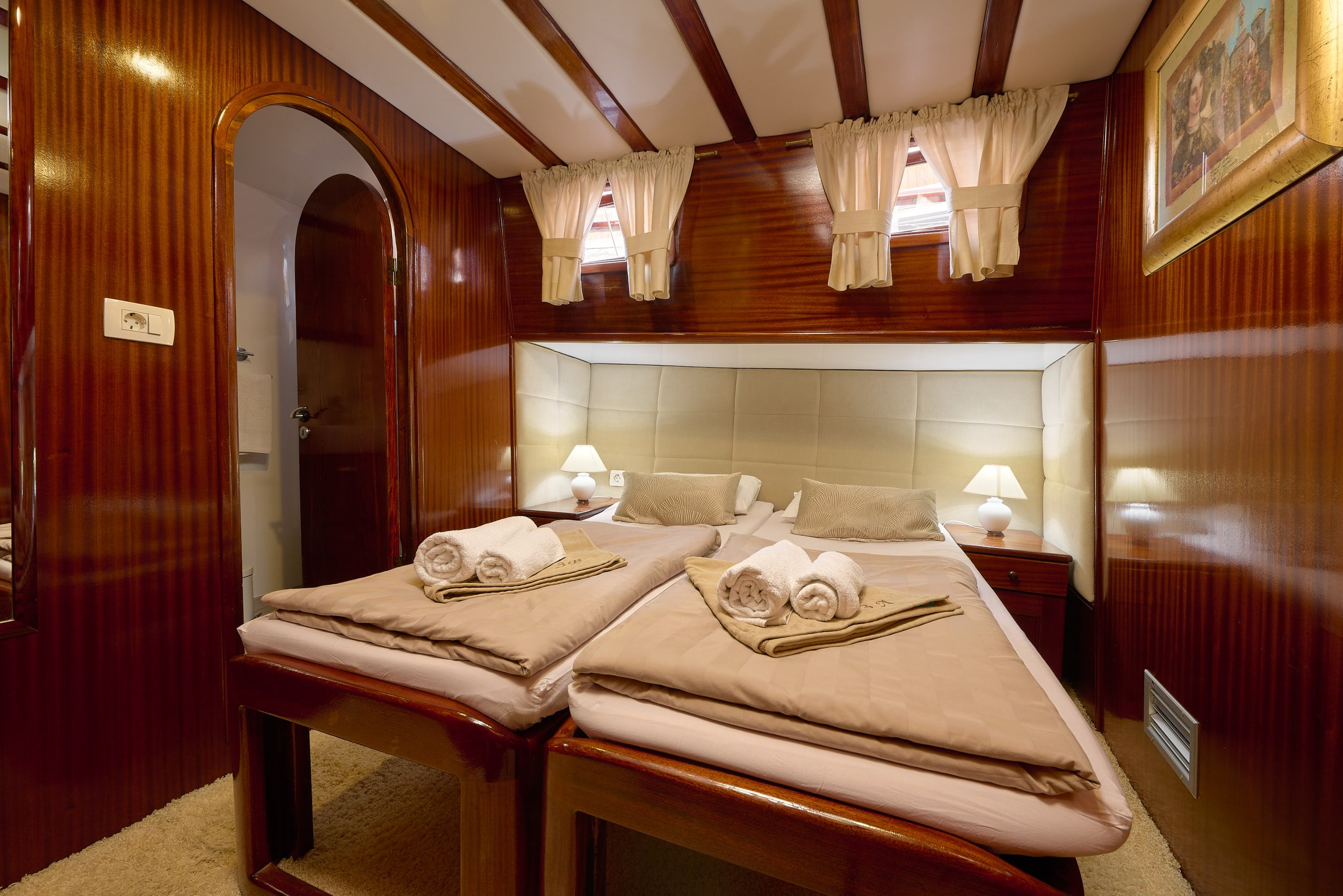 A cozy bedroom with twin beds, wooden walls, and beige bedding, with rolled towels on each bed, small lamps on nightstands, and small windows with curtains.