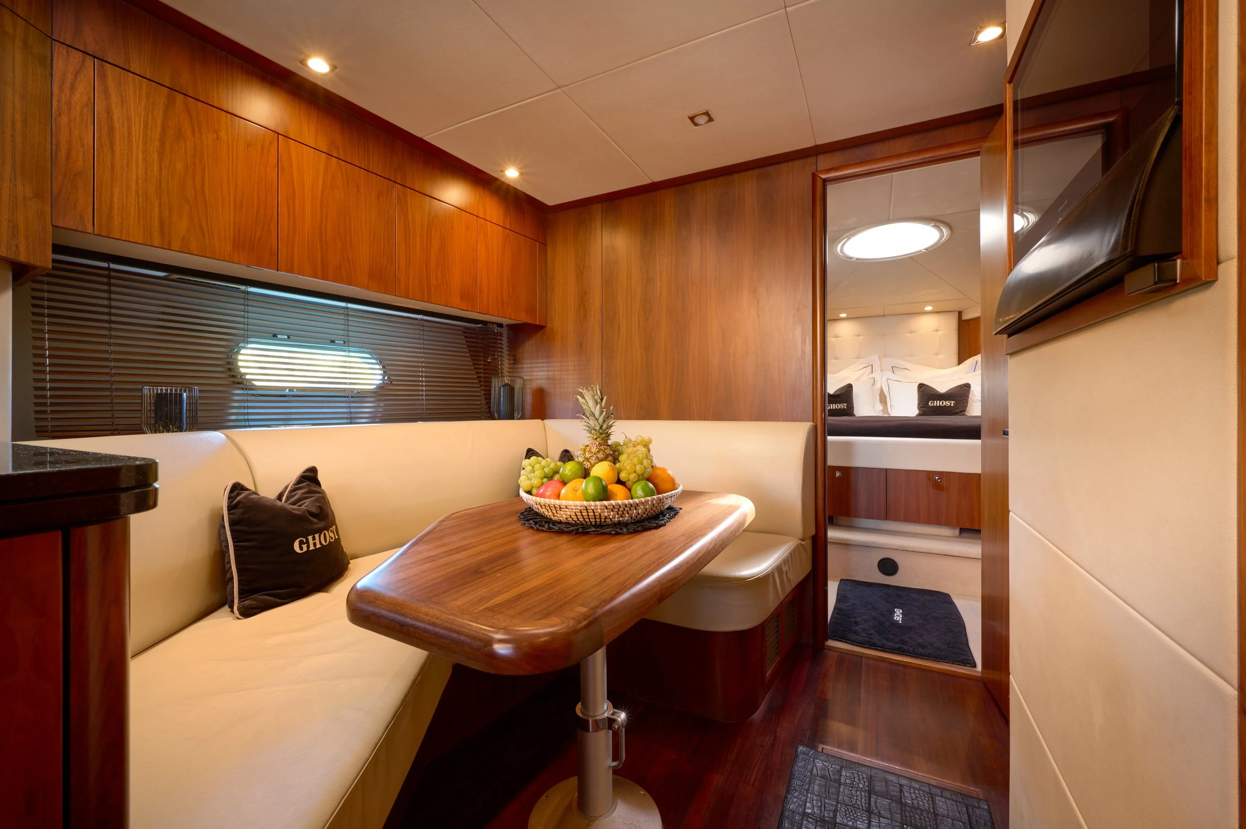 Interior of a yacht with a wooden dining table, cream cushioned seating, a bowl of mixed fruits, and a separate bedroom area with a bed, pillows, and soft lighting.