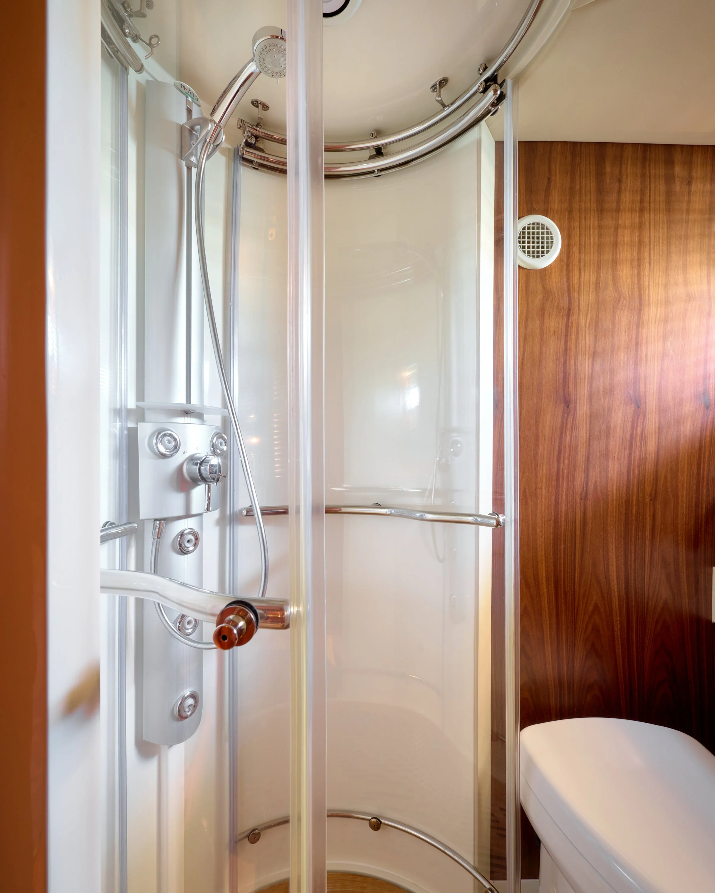 Inside a small bathroom with a shower stall with multiple shower heads and controls, a wooden wall, and a white toilet.