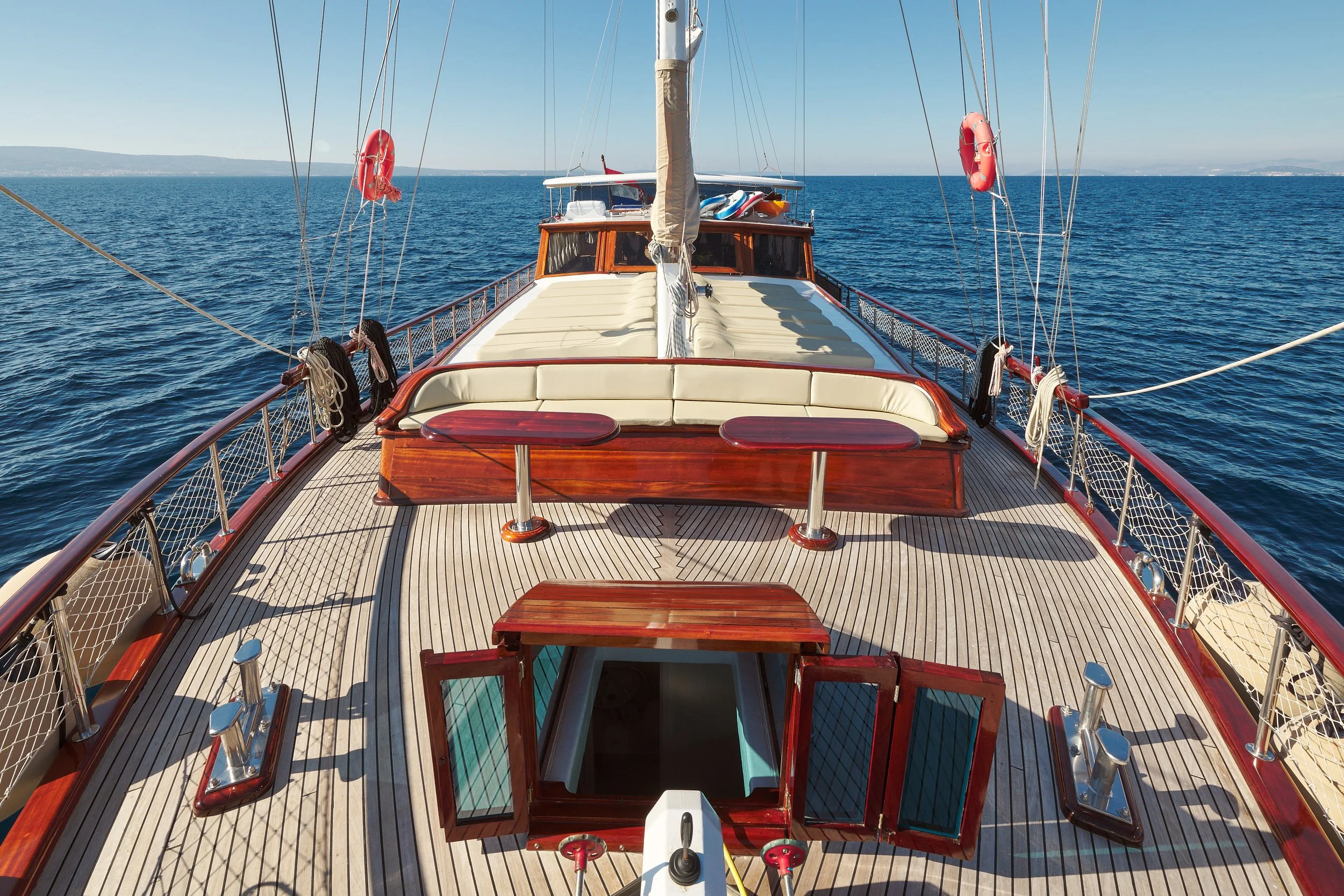 A view from the bow of a wooden sailing yacht on calm blue ocean waters, with a distant coastline on the horizon, clear blue sky, and some life preservers hanging on the boat.