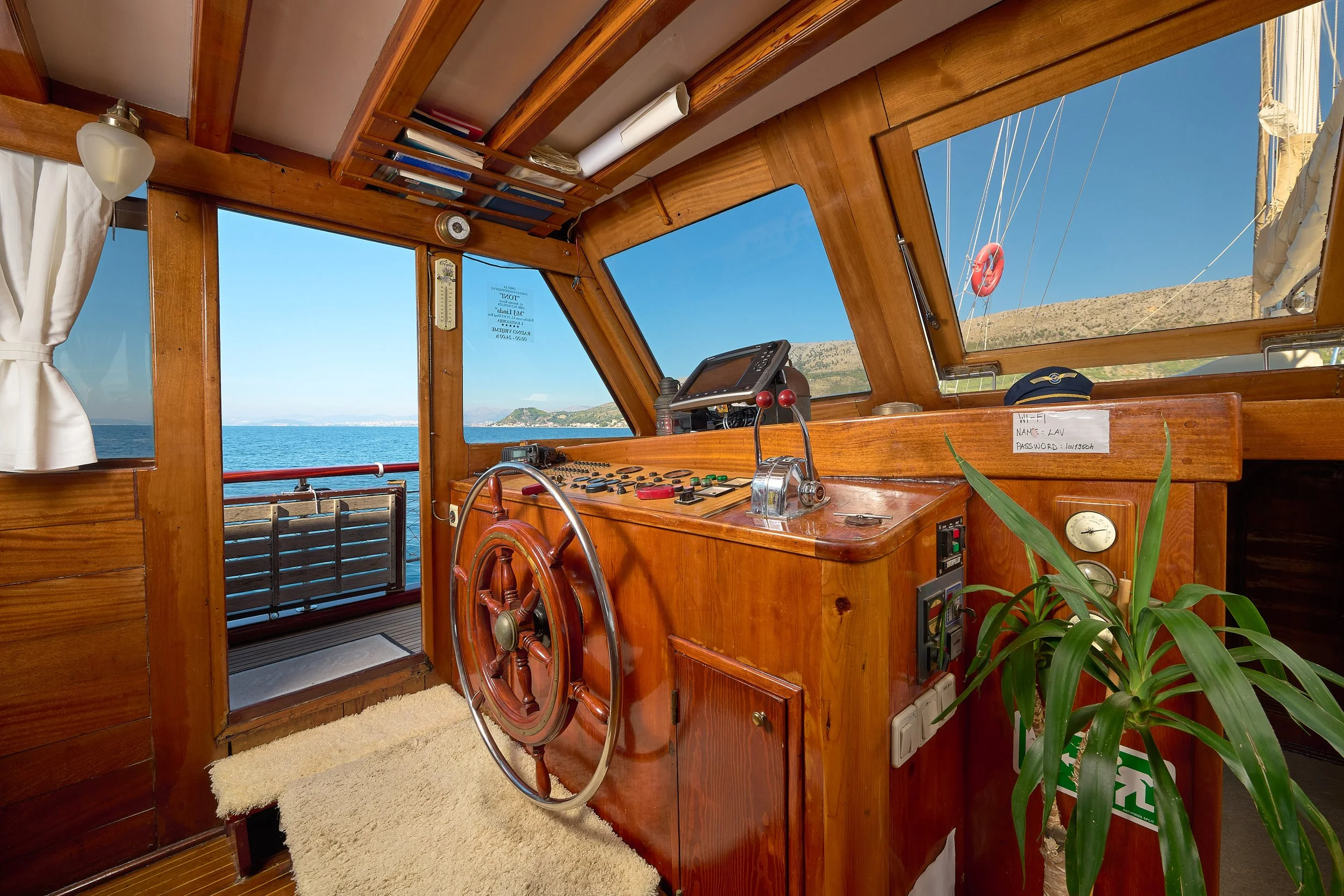 Inside a wooden boat's control room with sea view, steering wheel, navigation equipment, and a plant.