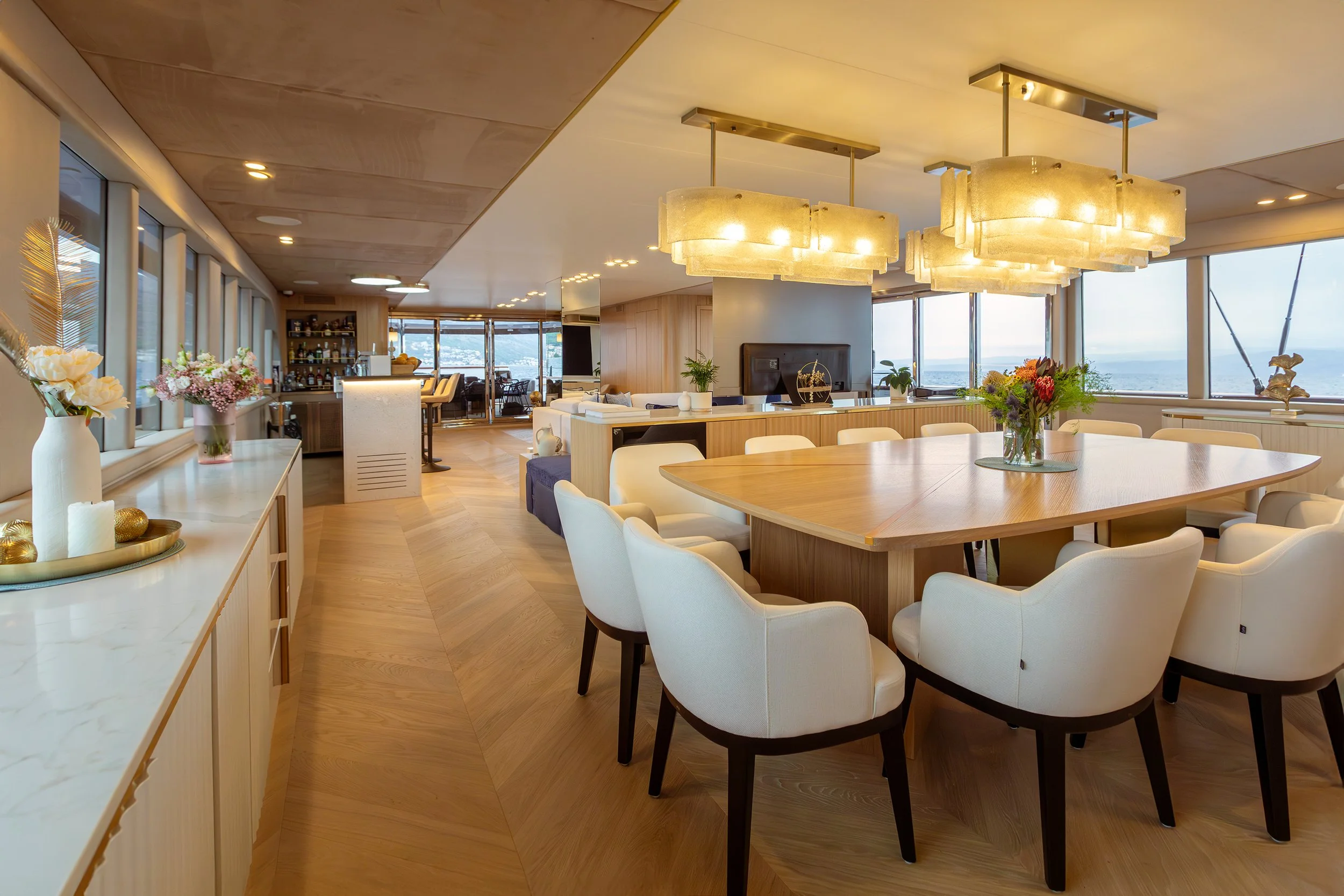 Elegant dining area with a large wooden table surrounded by white upholstered chairs, modern chandelier lights above, and large windows offering a view outside.