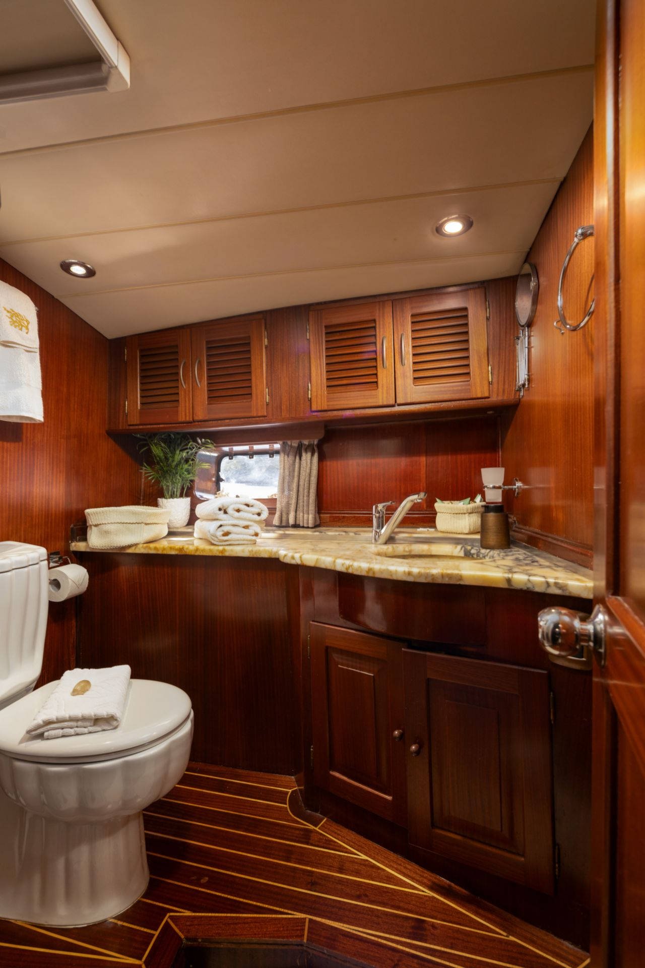Luxurious yacht bathroom with wooden cabinetry, marble countertop, toilet, towels, and porthole window.