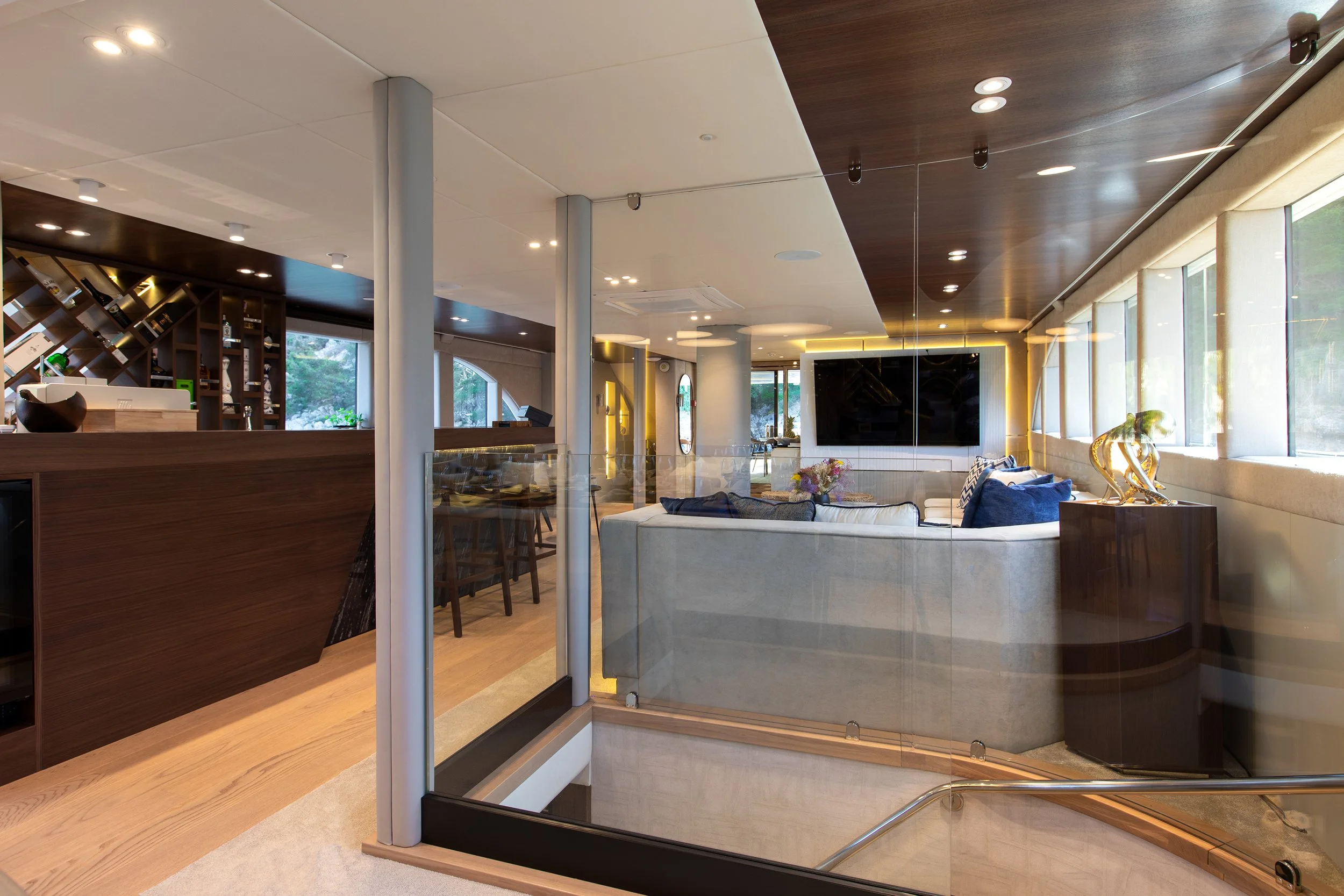 Modern living room with large windows, a curved sofa, a flat-screen TV on the wall, and decorative sculptures, with a view into the kitchen area behind a glass railing.