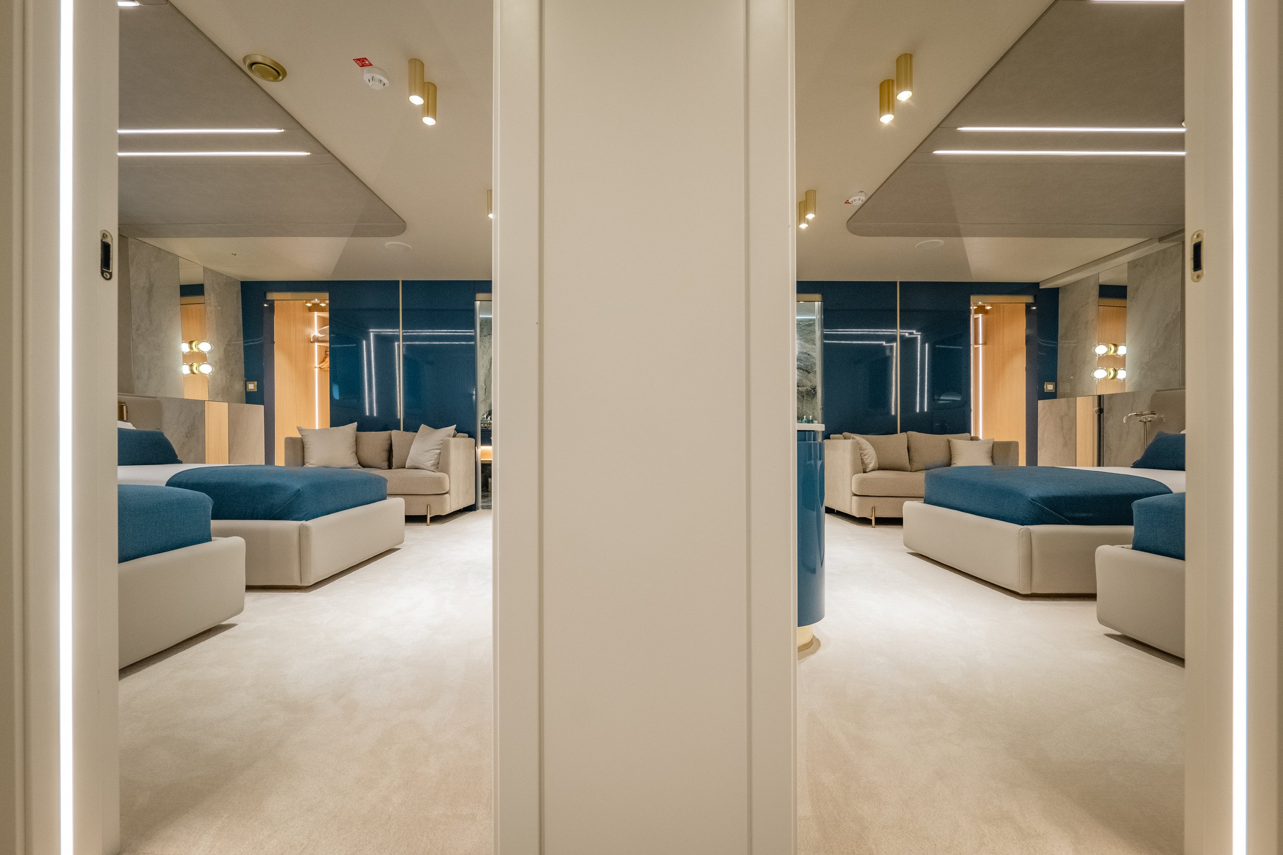 A hotel room with modern decor, featuring two large beds with blue bedding, beige sofas, and a navy accent wall. The room has contemporary lighting and mirrors, with a calm and elegant atmosphere.