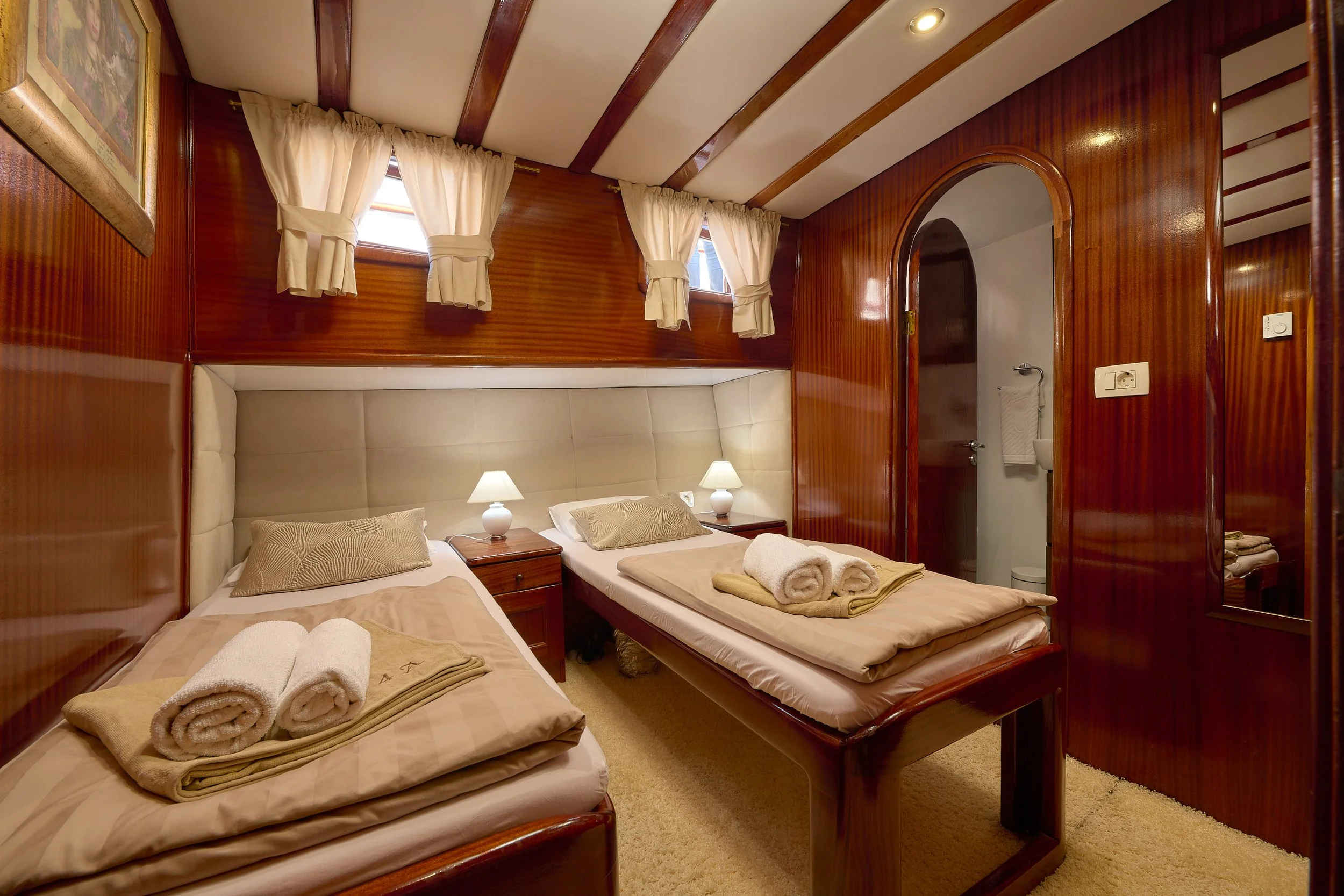 A cozy bedroom with two single beds featuring beige bedding and rolled towels on top. The room has wooden paneled walls, a beige upholstered headboard, and two small lamps on nightstands. There are three small windows with cream curtains, and a bathr