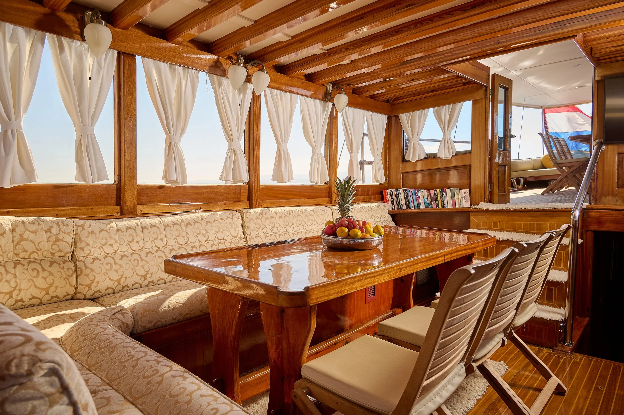 Interior of a wooden boat with a dining area, beige cushioned seating, a wood table with a fruit bowl, books, white curtains, and ocean view through windows.