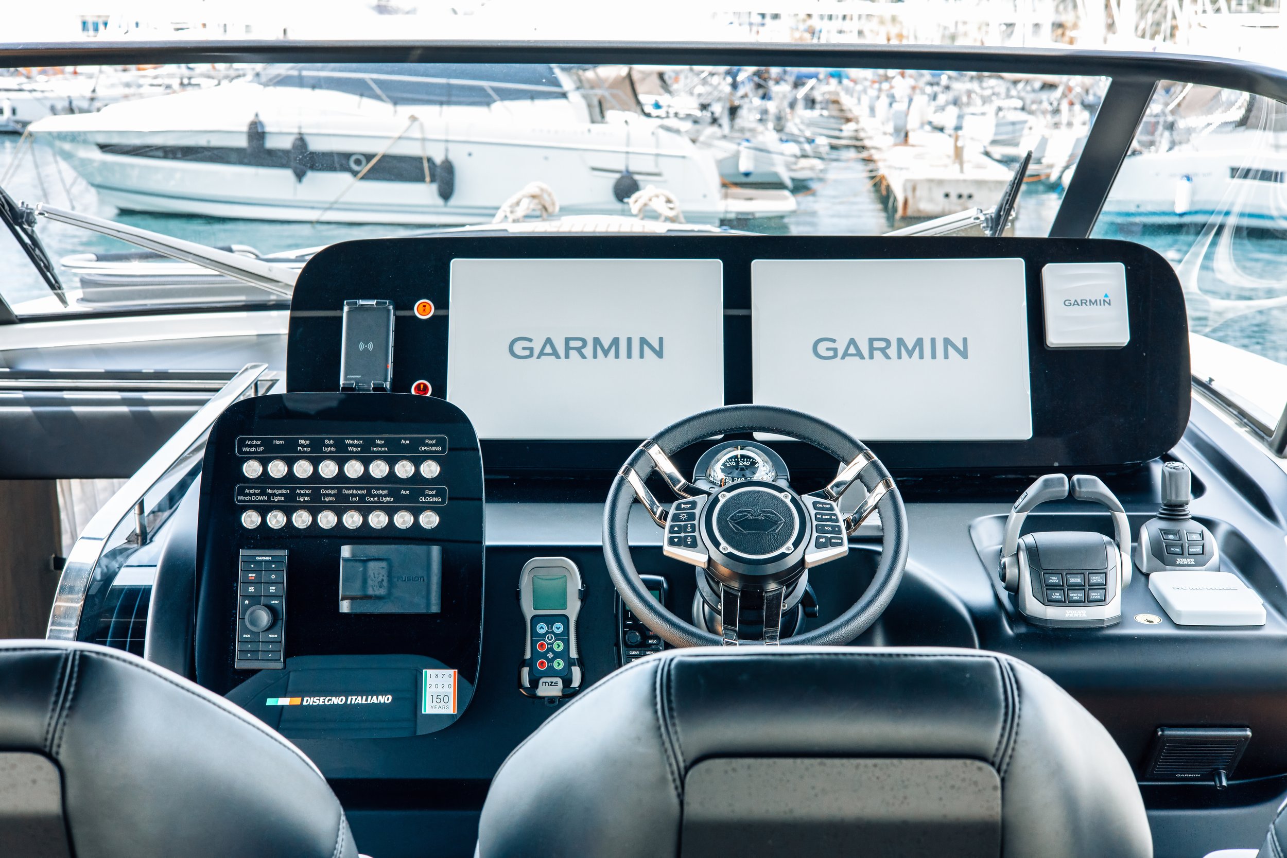 View of a yacht's control dashboard at a marina with boats docked in the background. The dashboard features two large Garmin screens, a steering wheel, and various controls and switches.