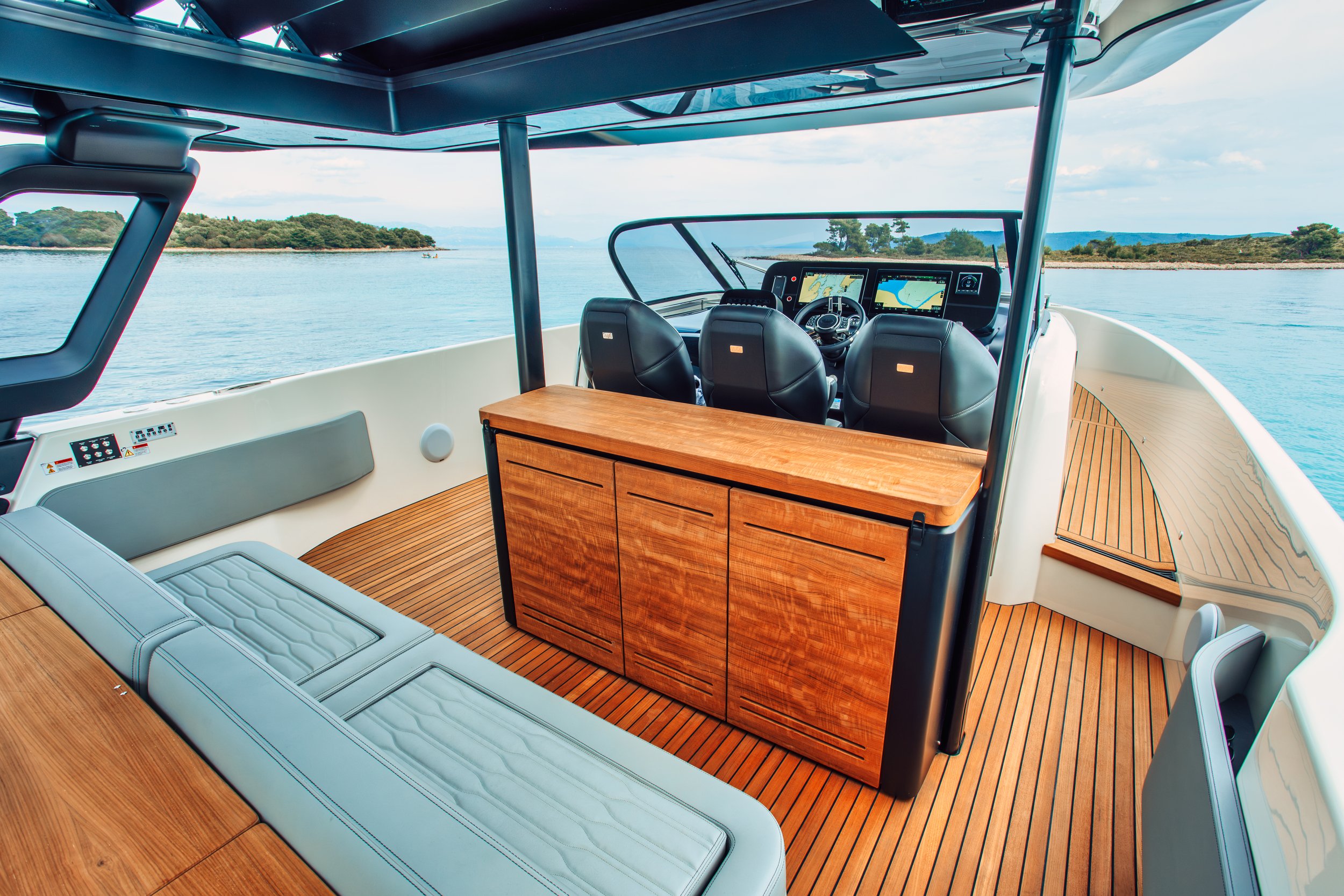 Interior of a boat with a wooden table, seating, and navigation screens, on water with a shoreline in the background.