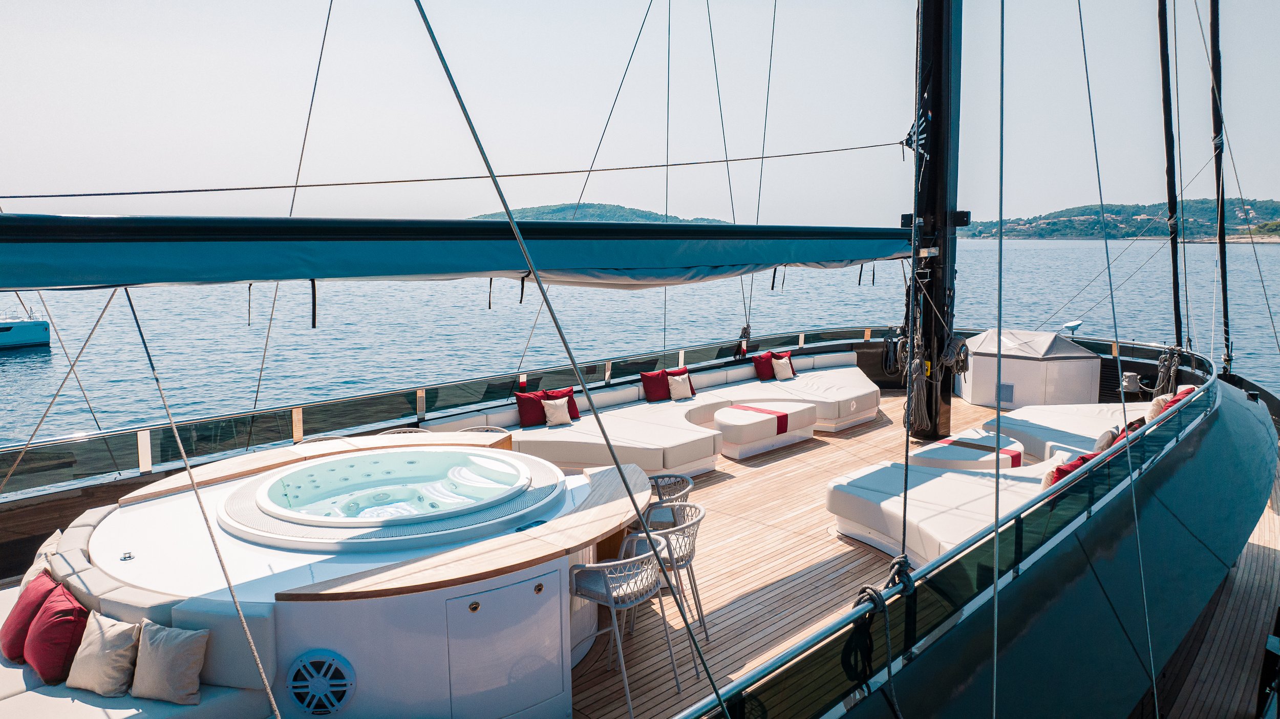 Luxury yacht with a hot tub and white seating area with red and beige cushions, docked on calm water with land in the background.