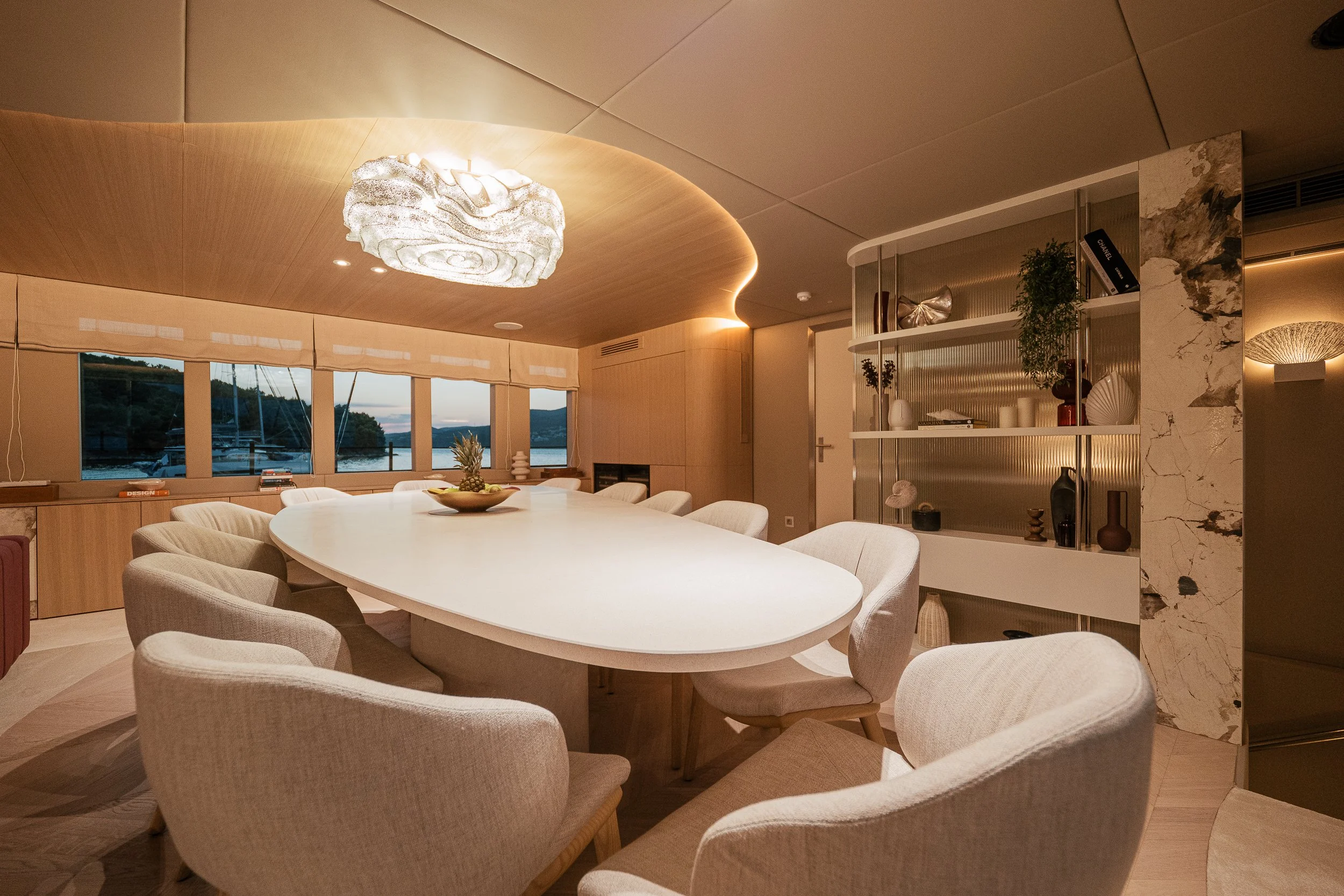 Modern dining room with an oval white table, surrounded by cream upholstered chairs, large windows with a view of a waterfront, decorative items on a curved shelf, and wooden ceiling and walls.