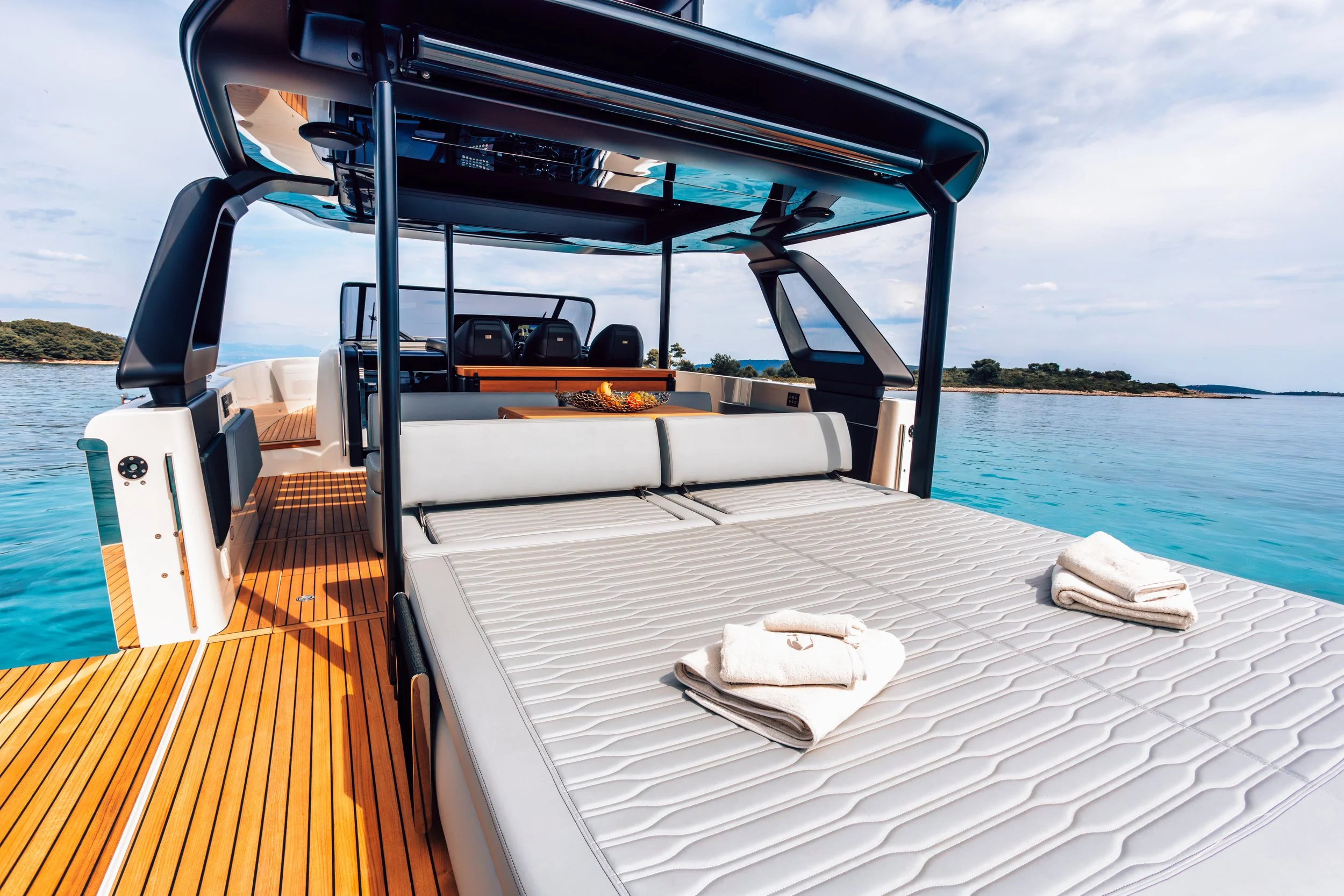 On a luxury yacht, a large sunbathing area with folded towels on a cushioned surface, overlooking calm blue water and distant shoreline under a partly cloudy sky.