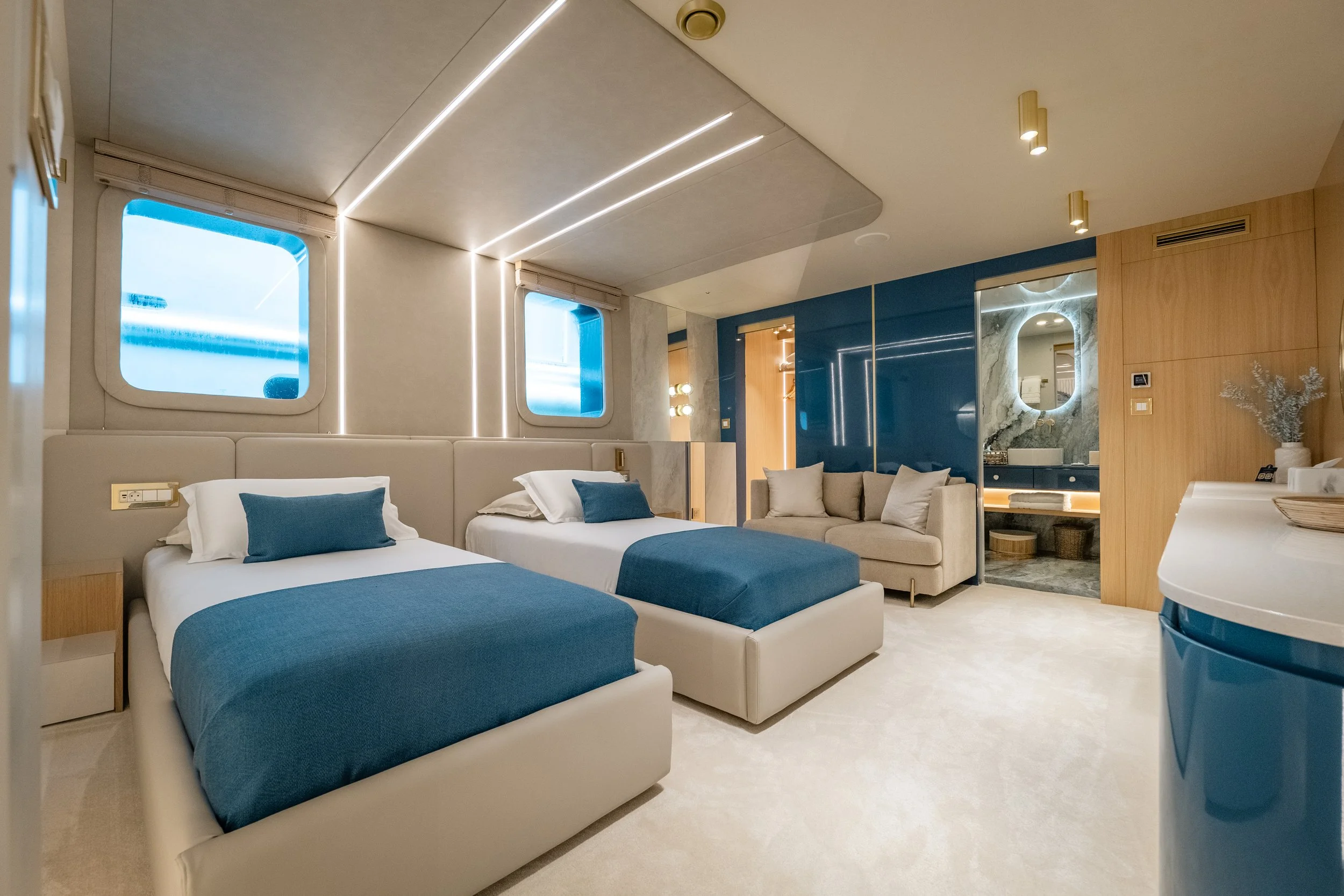 Modern hotel room with two beds, a beige sofa, and a bathroom with a round mirror