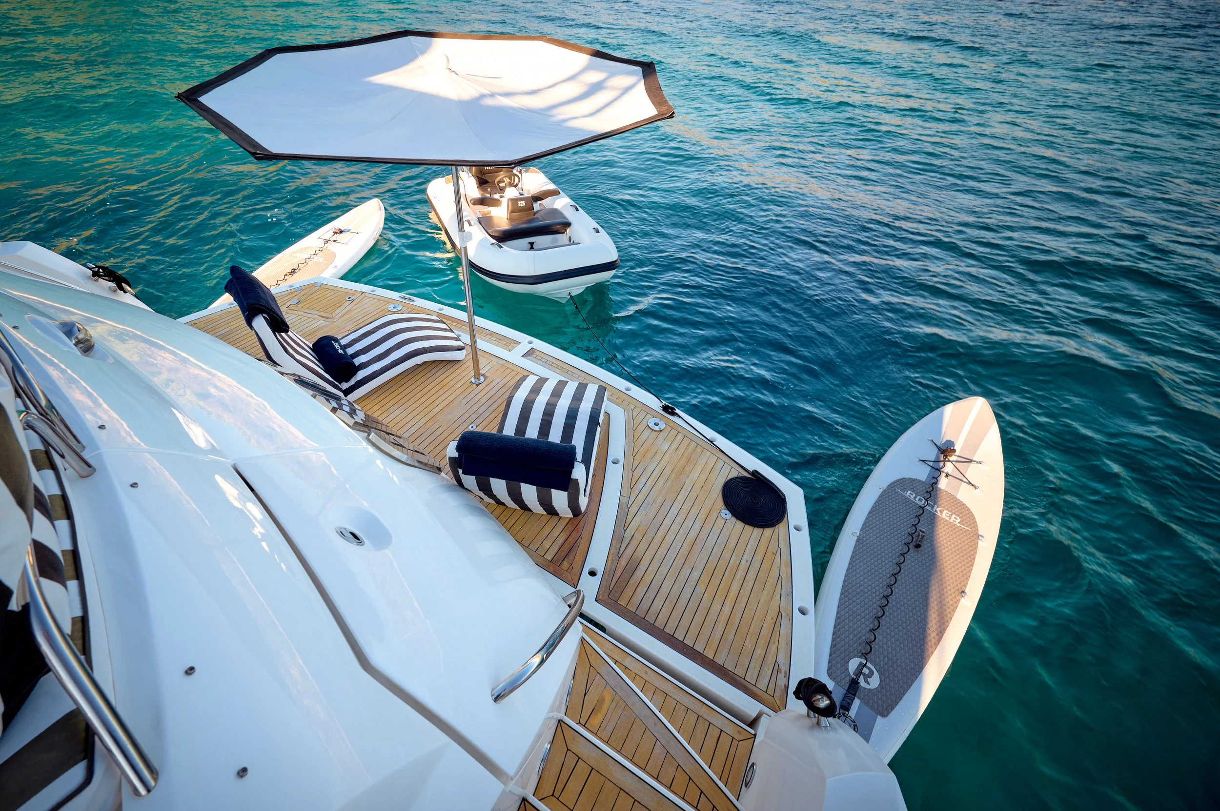 Aerial view of a yacht with a wooden deck, striped lounge chairs, and umbrellas, anchored in clear blue ocean water.