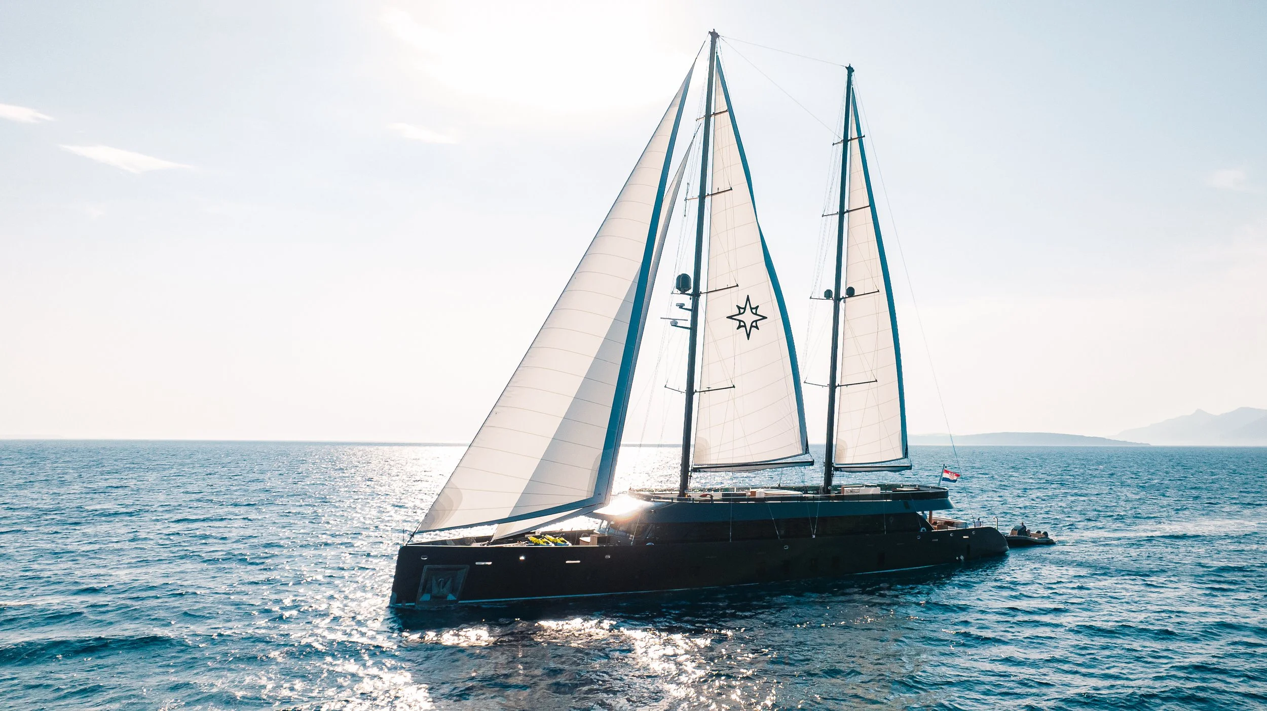 A large black sailing yacht with white sails on the ocean, reflecting sunlight, with a clear sky and distant mountains on the horizon.