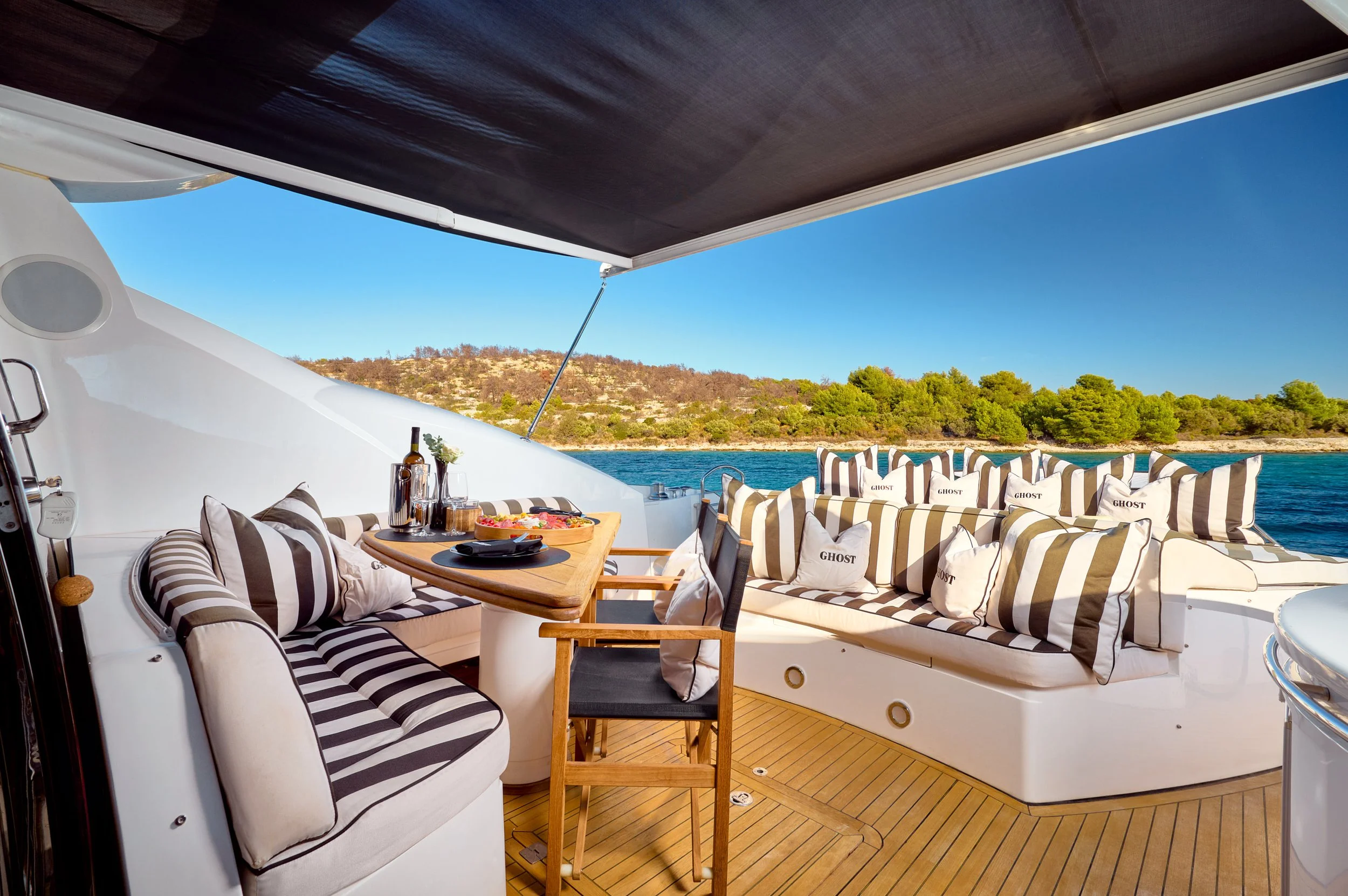 Luxury yacht deck with black and white striped cushions, a dining table set for a meal with wine and pizza, surrounded by chairs and cushions, overlooking a scenic water and green landscape under a blue sky.
