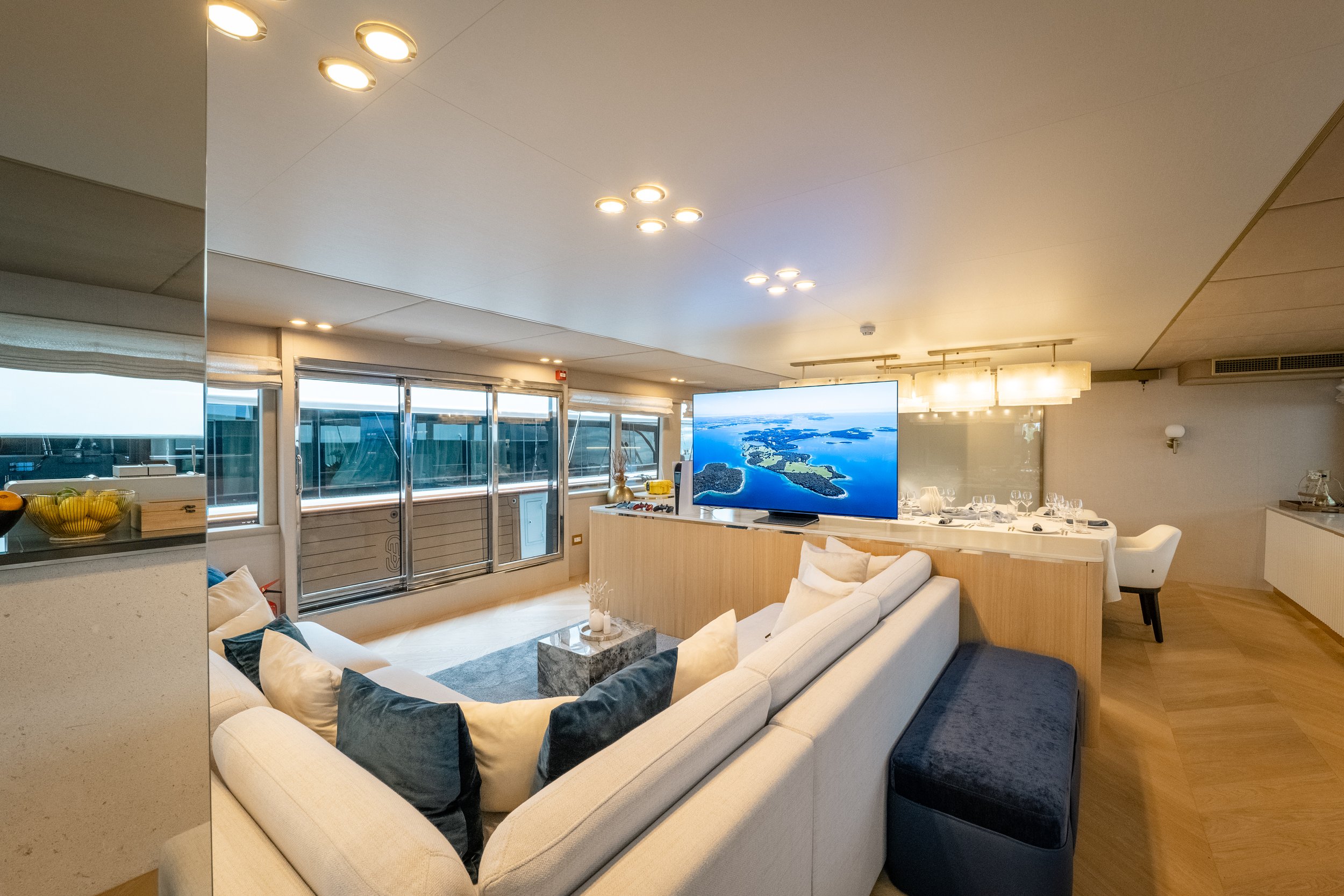 Open-concept living space with a large white sectional sofa, multiple cushions, a coffee table, a large TV displaying an aerial view of islands and water, and a dining area with a long table and several wine glasses. Large windows and sliding glass d