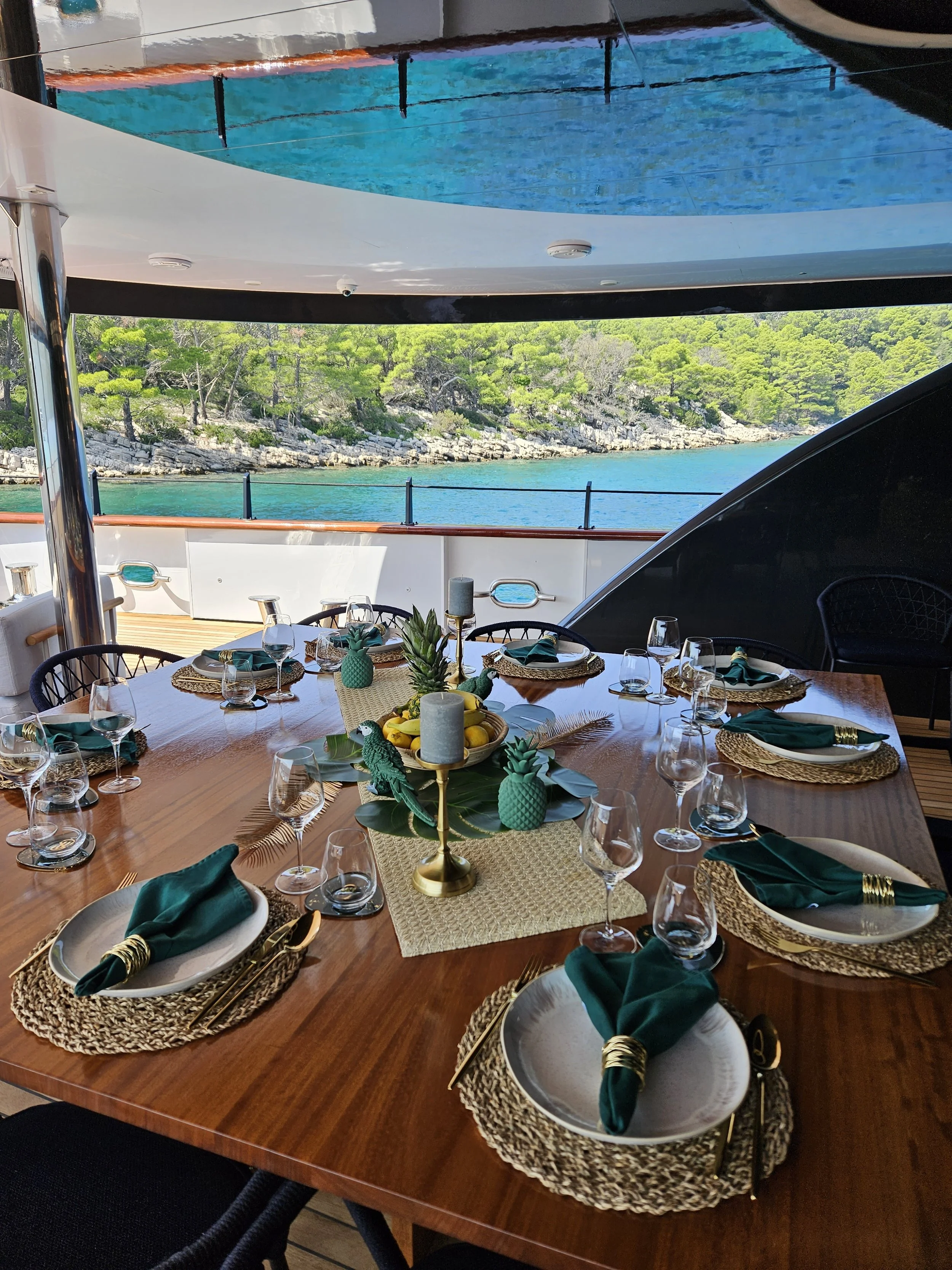 A dining table set with plates, green napkins, glassware, and tropical-themed centerpieces with pineapples and bananas, on a yacht overlooking the water.